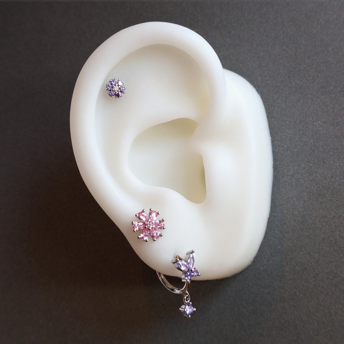 Nature-inspired silver ear piercing stack by JULE on a model, highlighting a large pink crystal flower stud, a purple butterfly dangle hoop, and a small purple floral helix piercing.