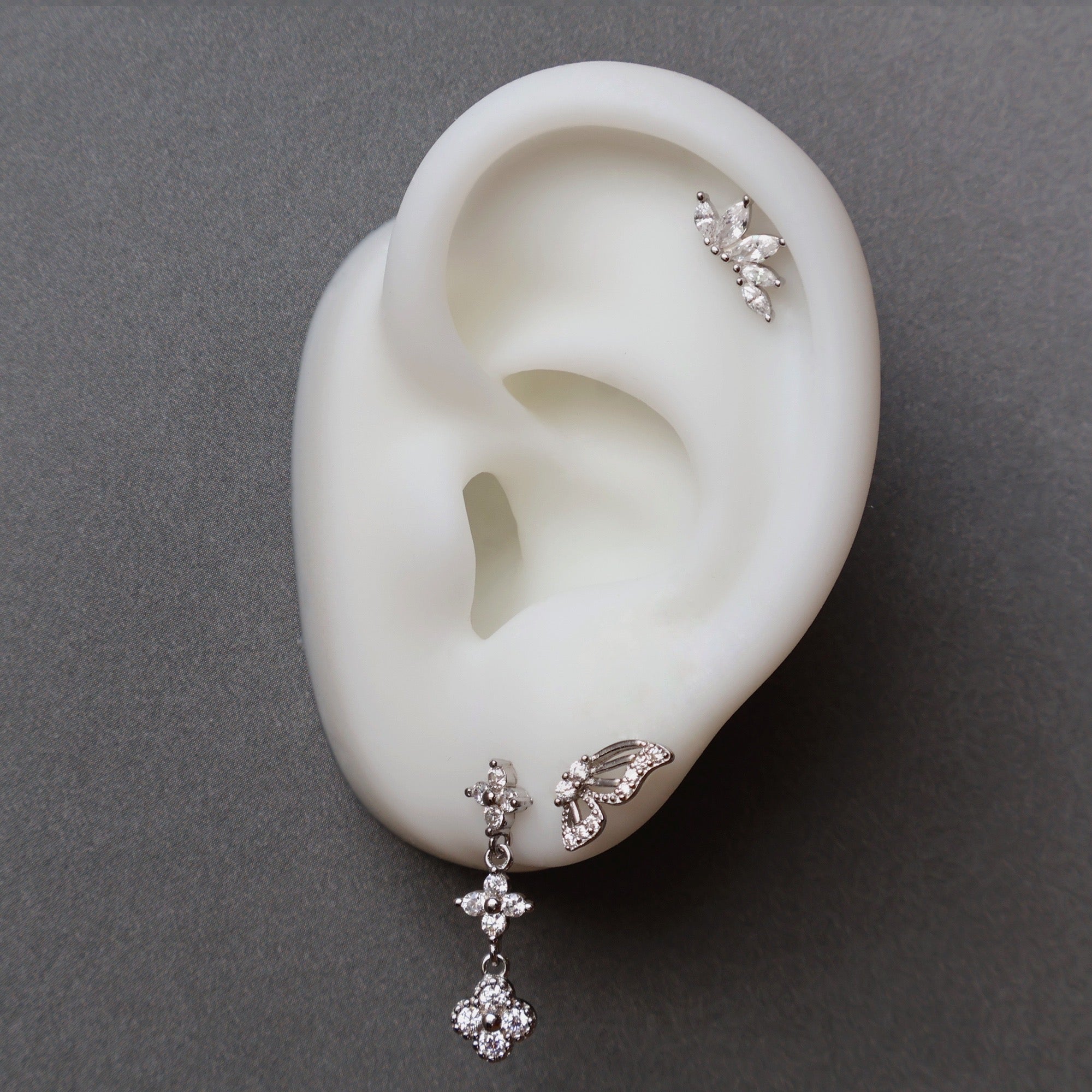 A close-up of a JULE ear piercing stack on a silicone ear, featuring a dangle floral stud, a butterfly wing, and a sparkling marquise leaf crawler on the cartilage.