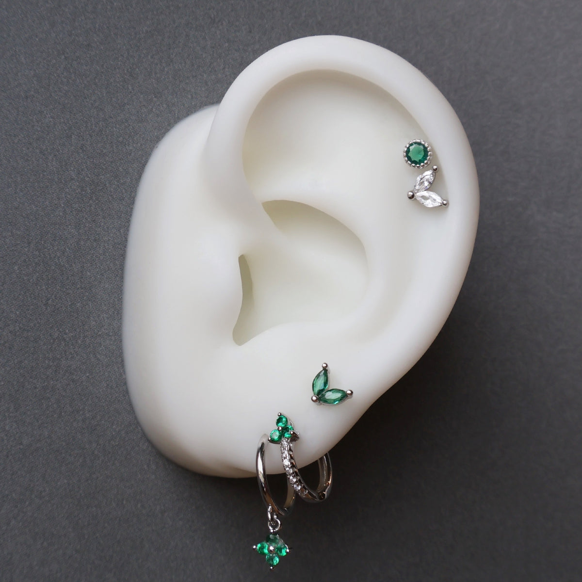 A curated silver ear piercing stack on a white display model by JULE, featuring a green clover dangle hoop, a paved huggie, a green leaf stud on the upper lobe, and a double helix piercing with round and leaf-shaped studs.