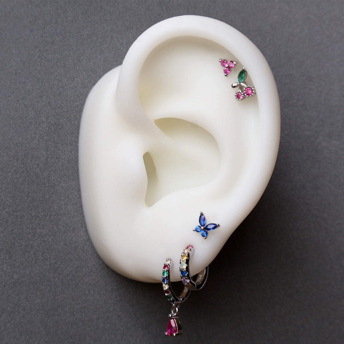 Silver ear piercing stack on a model featuring a multi-colored huggie with a red dangle, a blue butterfly stud on the lobe, along with a cherry stud and pink gemstone cluster on the helix by JULE.