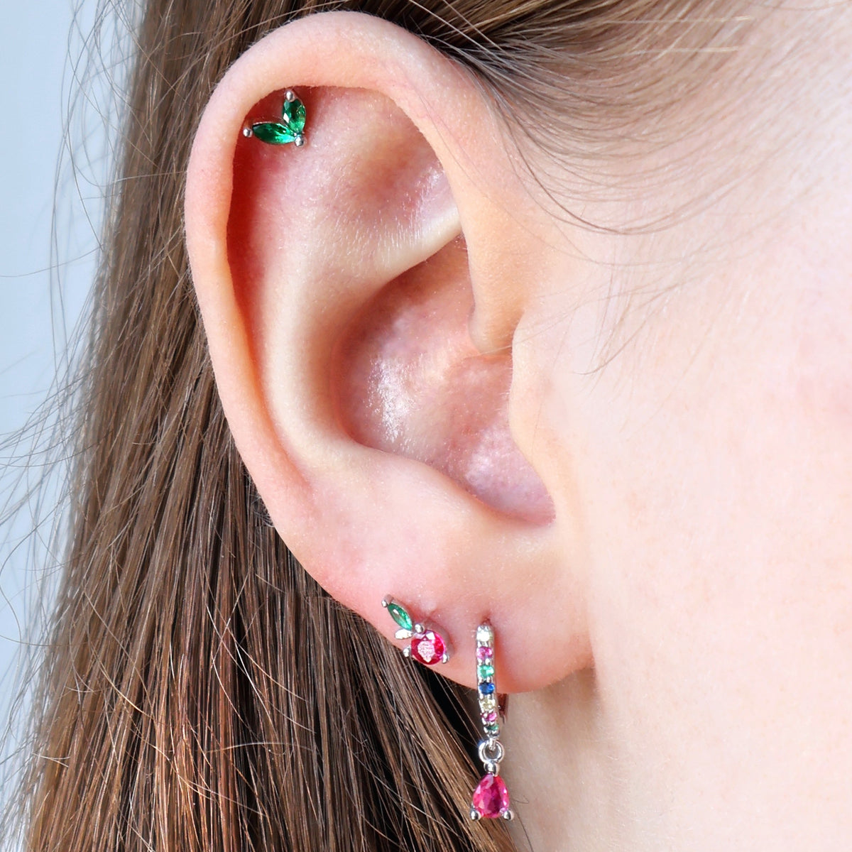 A curated silver ear piercing stack on a real ear, showcasing a sparkling cherry stud, a colorful gemstone huggie with a red teardrop dangle on the lobe, and a green leaf piercing on the helix by JULE.
