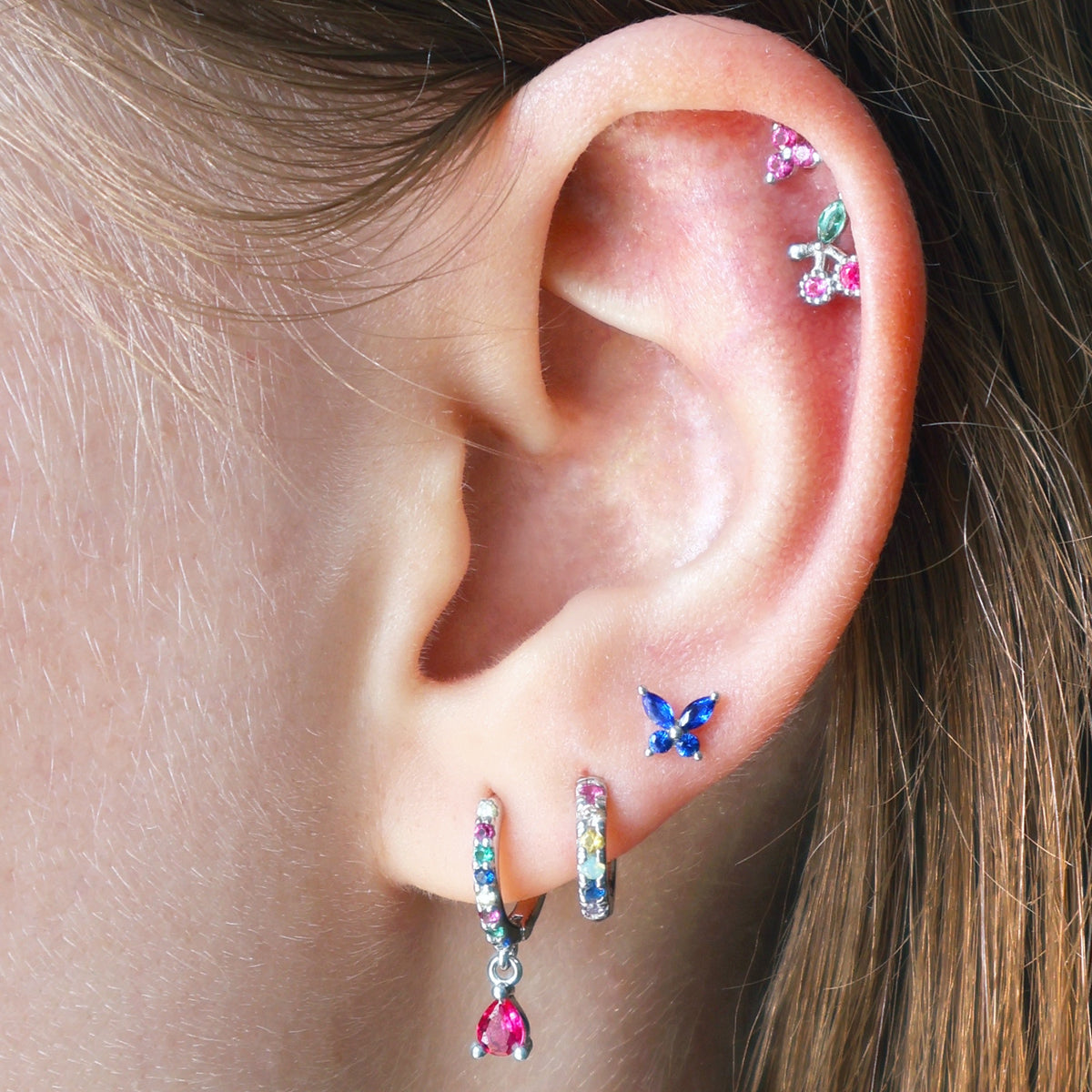 A stunning multi-piercing ear stack by JULE worn on an ear, featuring two colorful gemstone huggies, a blue butterfly stud on the lobe, and a vibrant double helix with a cherry and pink crystal cluster.