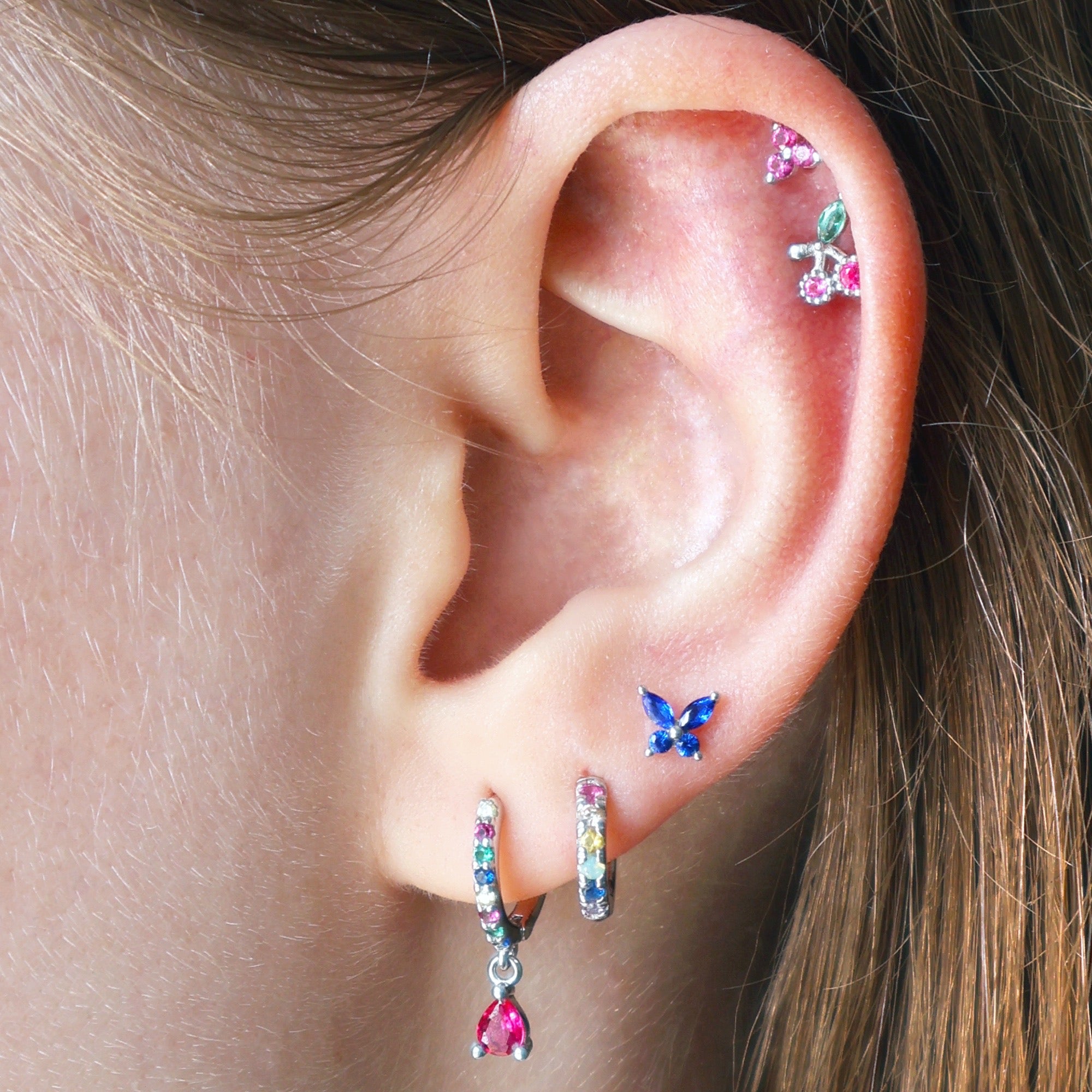 A stunning multi-piercing ear stack by JULE worn on an ear, featuring two colorful gemstone huggies, a blue butterfly stud on the lobe, and a vibrant double helix with a cherry and pink crystal cluster.