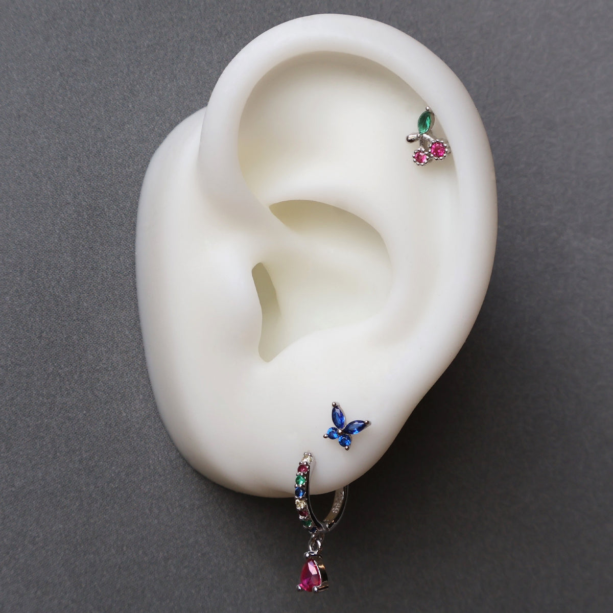 A vibrant silver ear piercing stack on a display model by JULE, featuring a colorful gemstone huggie with a red teardrop, a blue butterfly stud on the lobe, and a sparkling cherry piercing on the helix.