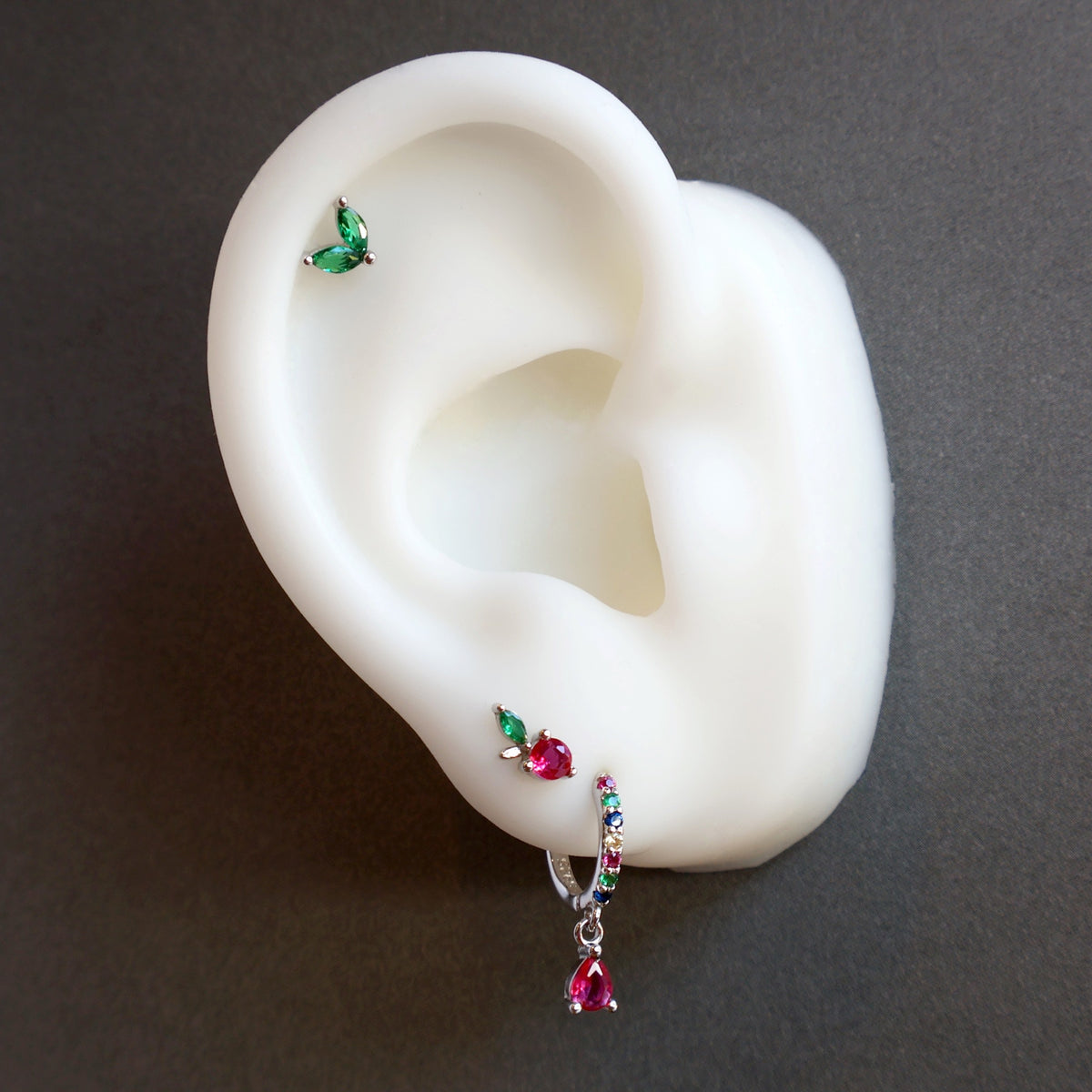 A delicate silver ear piercing stack on a display model, highlighting a colorful huggie with a red teardrop, a small fruit stud with a green leaf on the lobe, and a double green leaf helix piercing.
