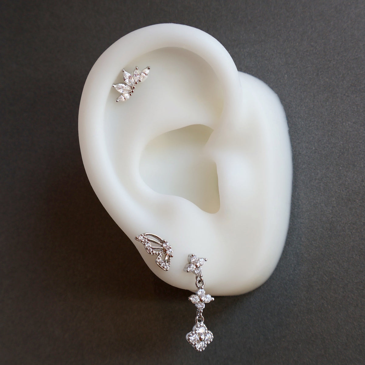 A curated silver ear piercing stack on a white display model by JULE, featuring a floral dangle stud on the lobe, a sparkling butterfly wing, and a crystal leaf helix piercing.
