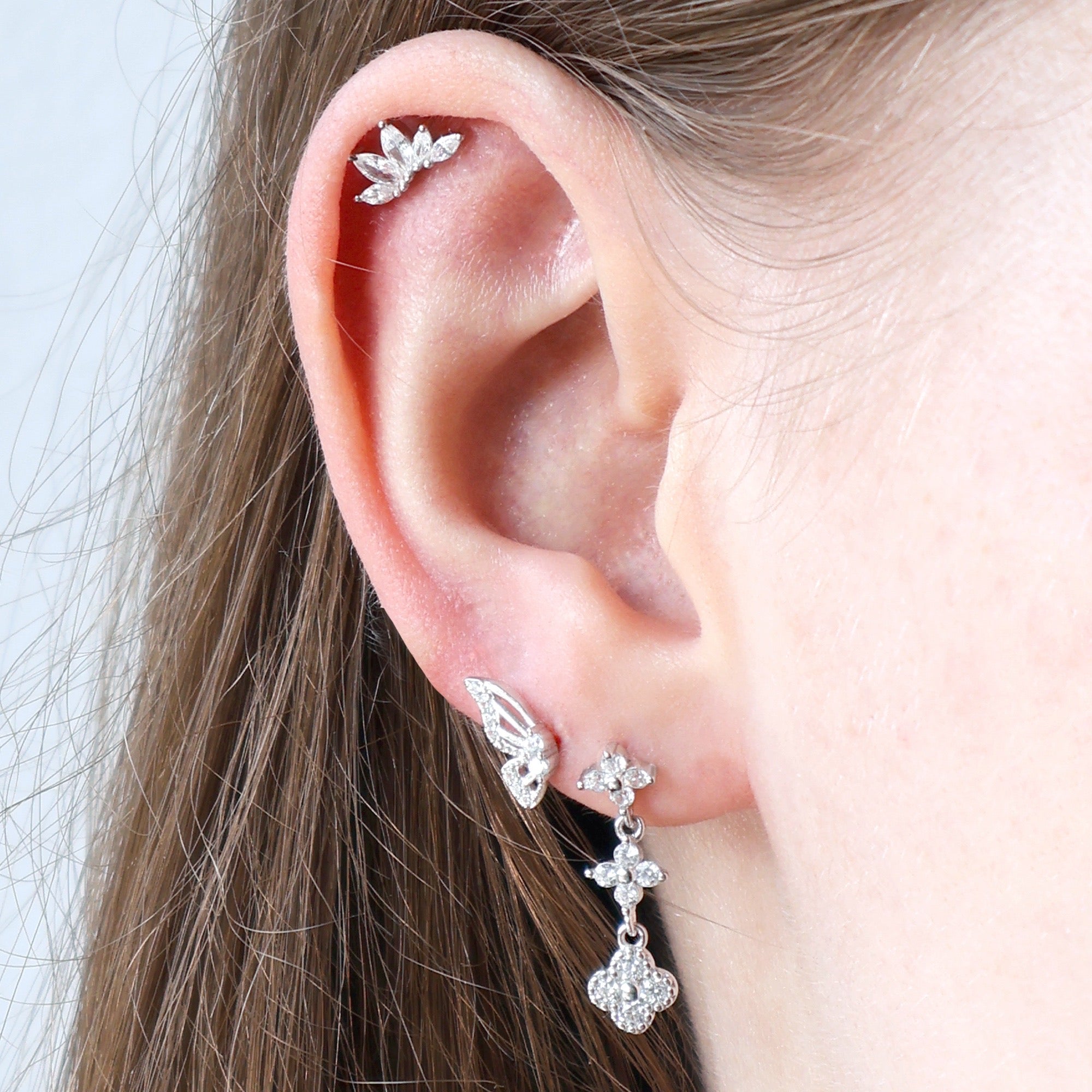Close-up of a woman's ear adorned with a JULE silver piercing stack, including a sparkling floral dangle stud, a butterfly wing, and a crystal marquise leaf helix piercing.