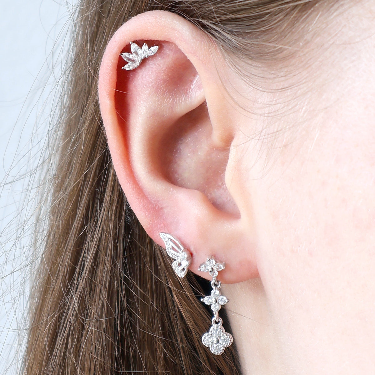 Close-up of a woman's ear adorned with a JULE silver piercing stack, including a sparkling floral dangle stud, a butterfly wing, and a crystal marquise leaf helix piercing.
