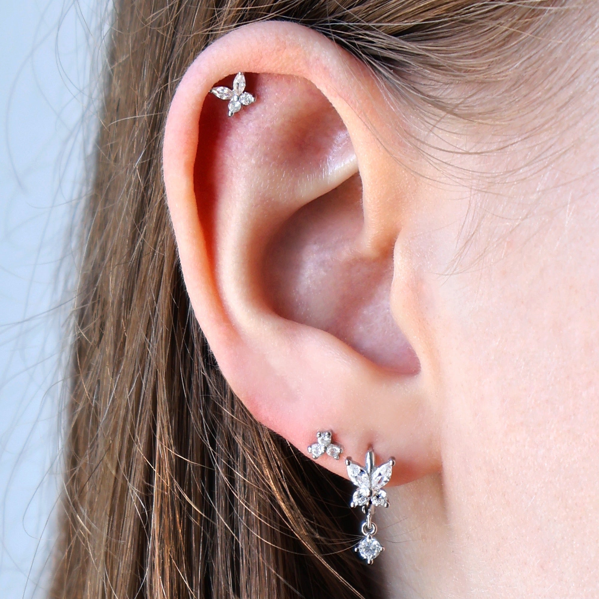 A woman wearing a beautiful silver ear piercing stack by JULE, featuring a clear crystal butterfly dangle hoop, a delicate cluster stud on the lobe, and a matching crystal butterfly piercing on the helix.