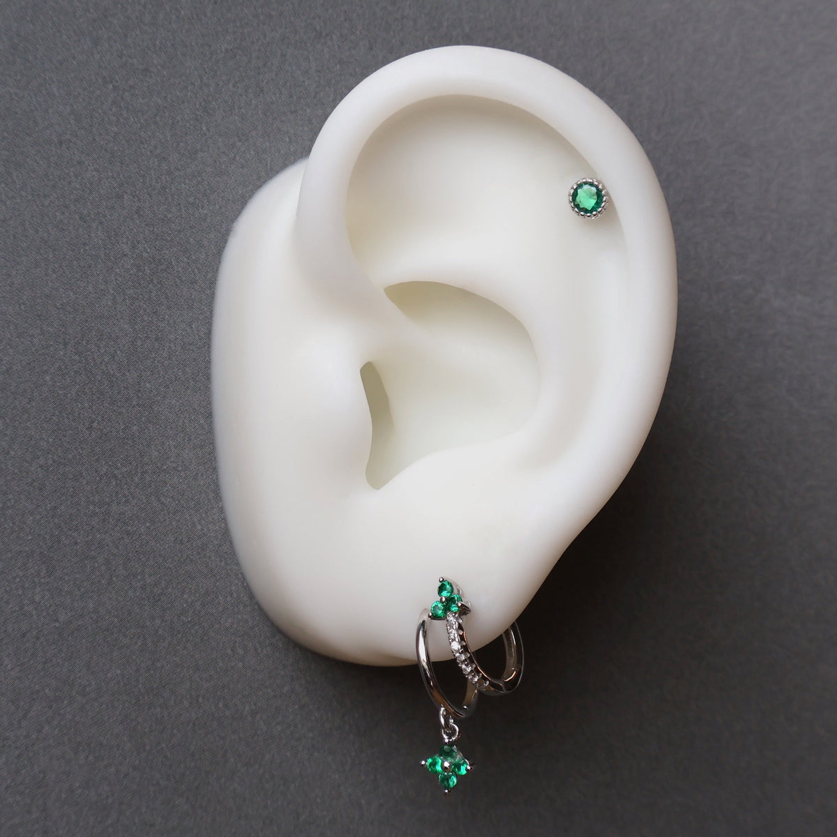 A styled ear piercing stack by JULE featuring a green gemstone dangle hoop on the first lobe, a matching green huggie on the second lobe, and a round green stud on the helix.