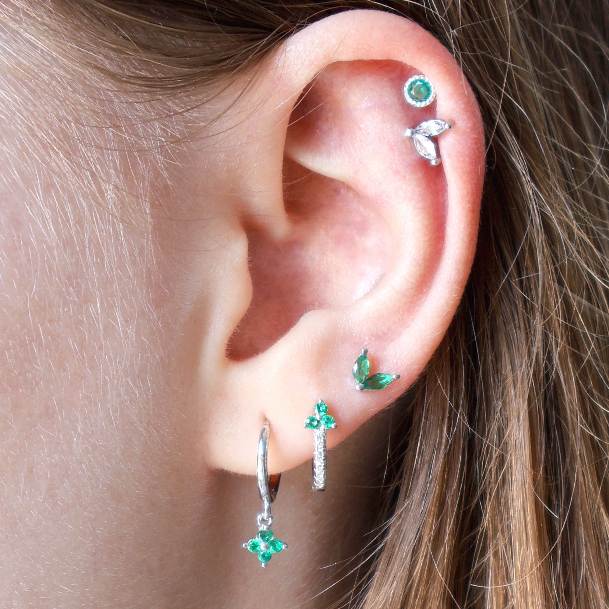 A woman wearing a nature-inspired silver ear piercing stack by JULE, showcasing green gemstone hoops on the lobes, a delicate green leaf stud, and a beautiful double helix piercing.