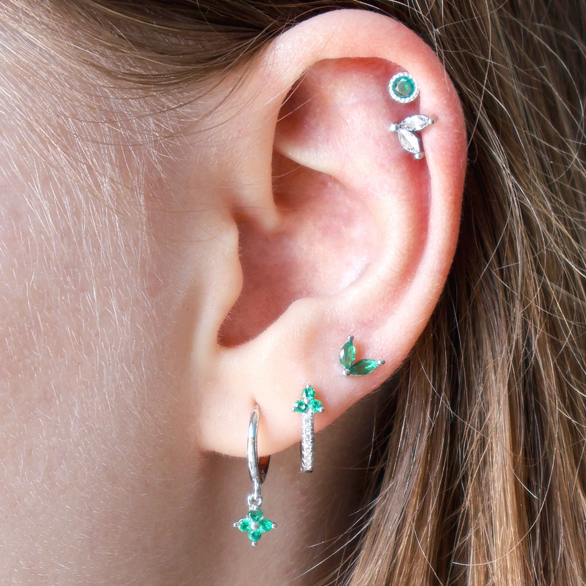 A woman wearing a nature-inspired silver ear piercing stack by JULE, showcasing green gemstone hoops on the lobes, a delicate green leaf stud, and a beautiful double helix piercing.