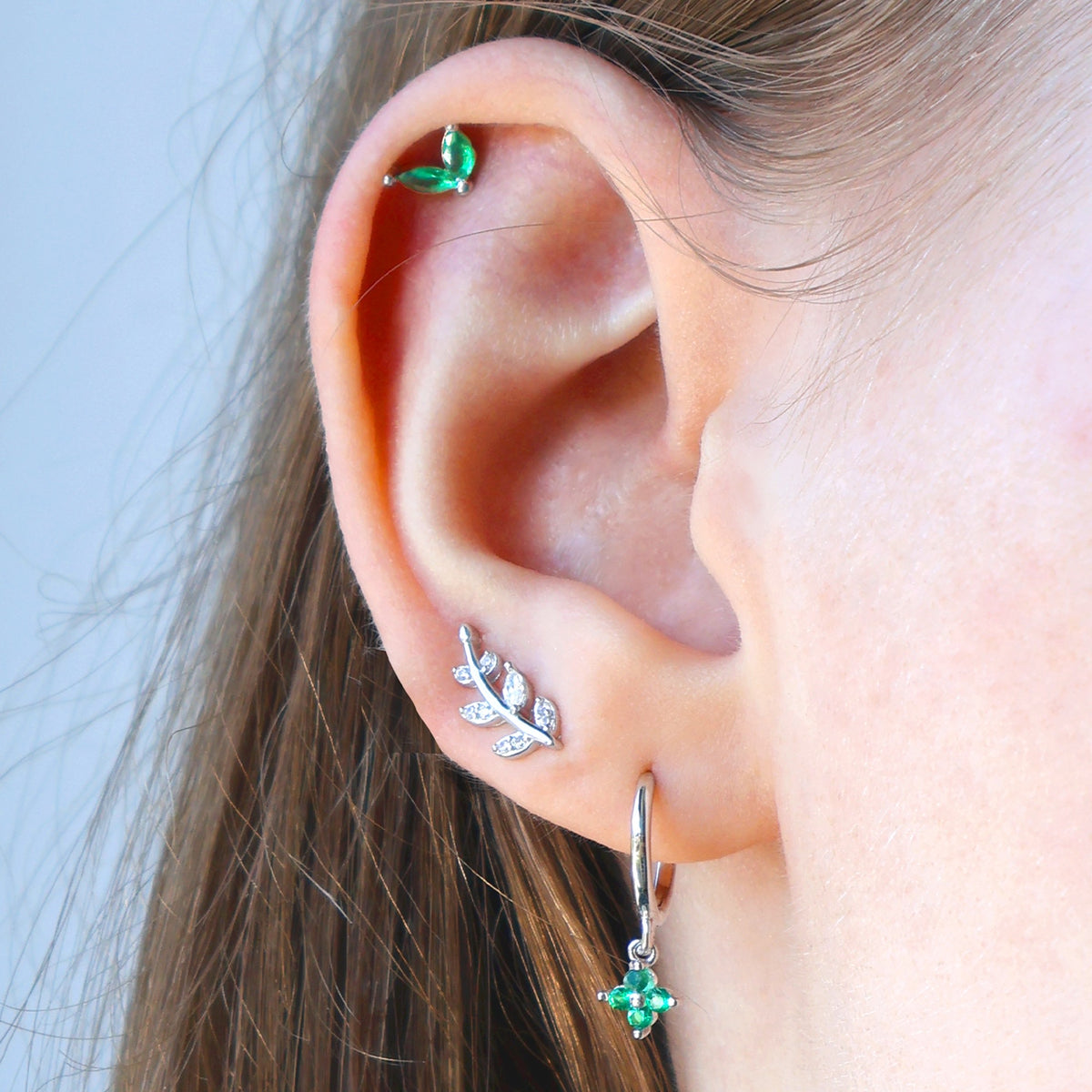 A woman wearing a nature-inspired JULE ear piercing stack with a green gemstone dangle hoop, a silver leaf crawler on the second lobe, and a green leaf stud on the helix.