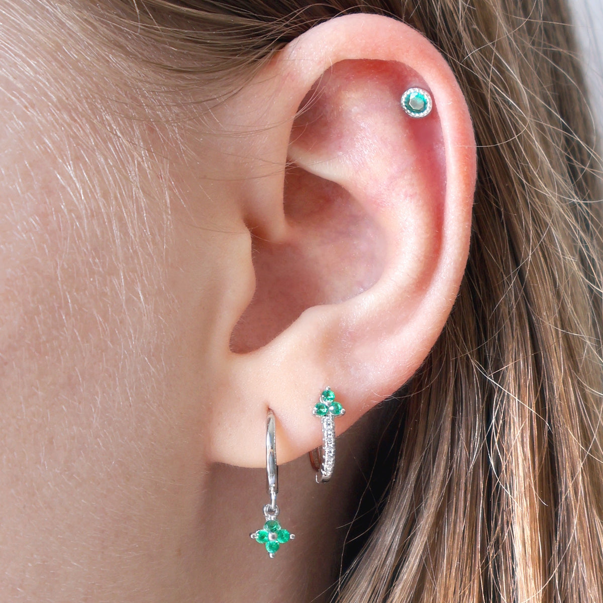 A woman wearing ear piercing stack by JULE featuring a green gemstone dangle hoop on the first lobe, a matching green huggie on the second lobe, and a round green stud on the helix.