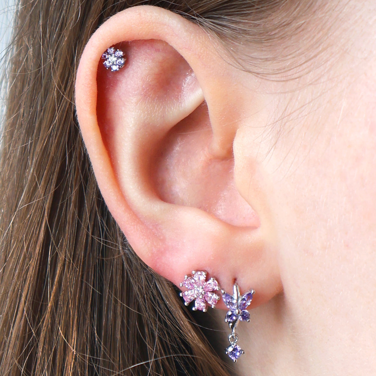 Close-up of a styled silver ear piercing stack on a real ear by JULE, showcasing a vibrant pink crystal flower stud on the lobe, a purple butterfly dangle, and a delicate purple floral helix piercing.