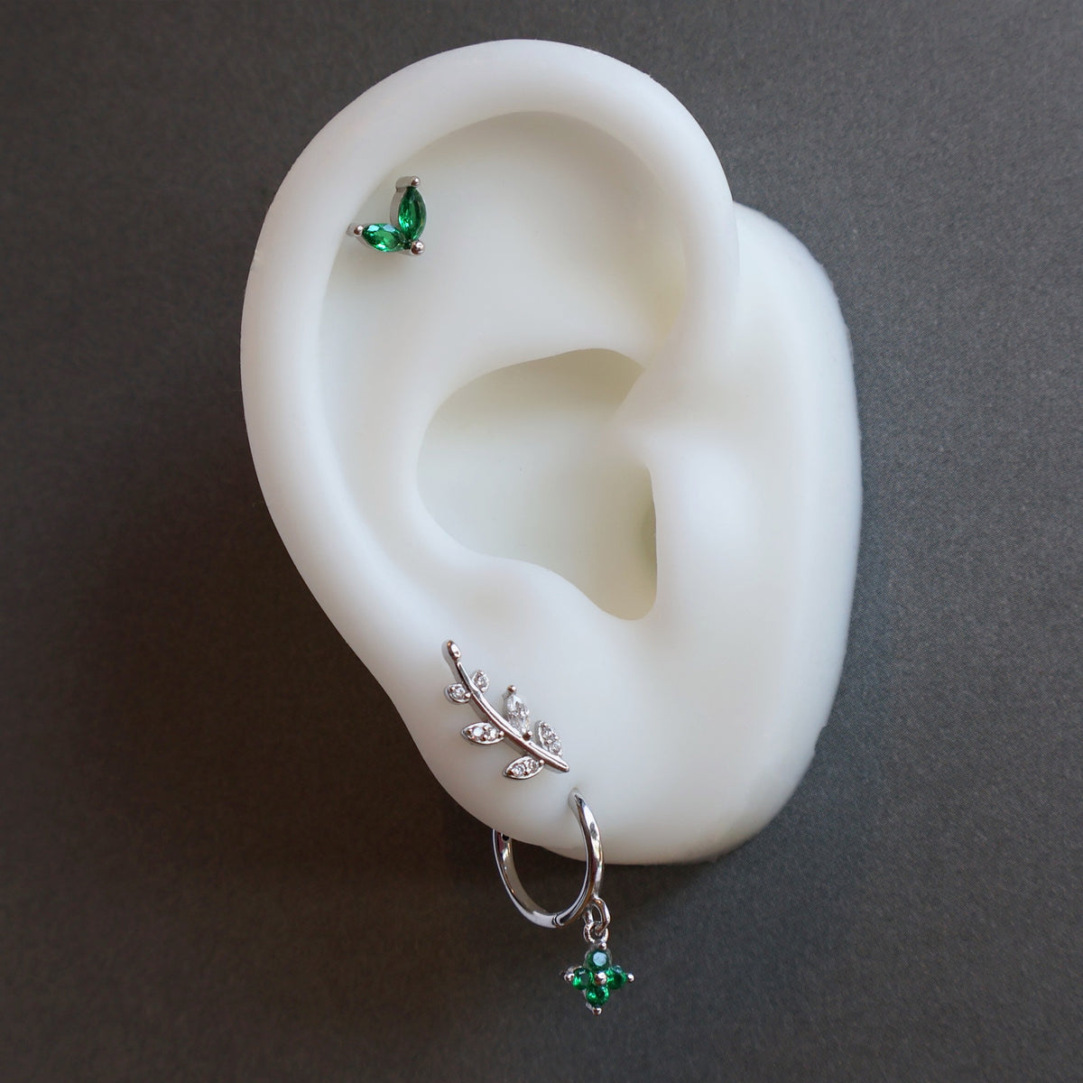 Close-up of a JULE ear piercing stack on a silicone model, showcasing a green clover dangle hoop on the lobe, a silver leaf climber, and a green leaf helix stud.
