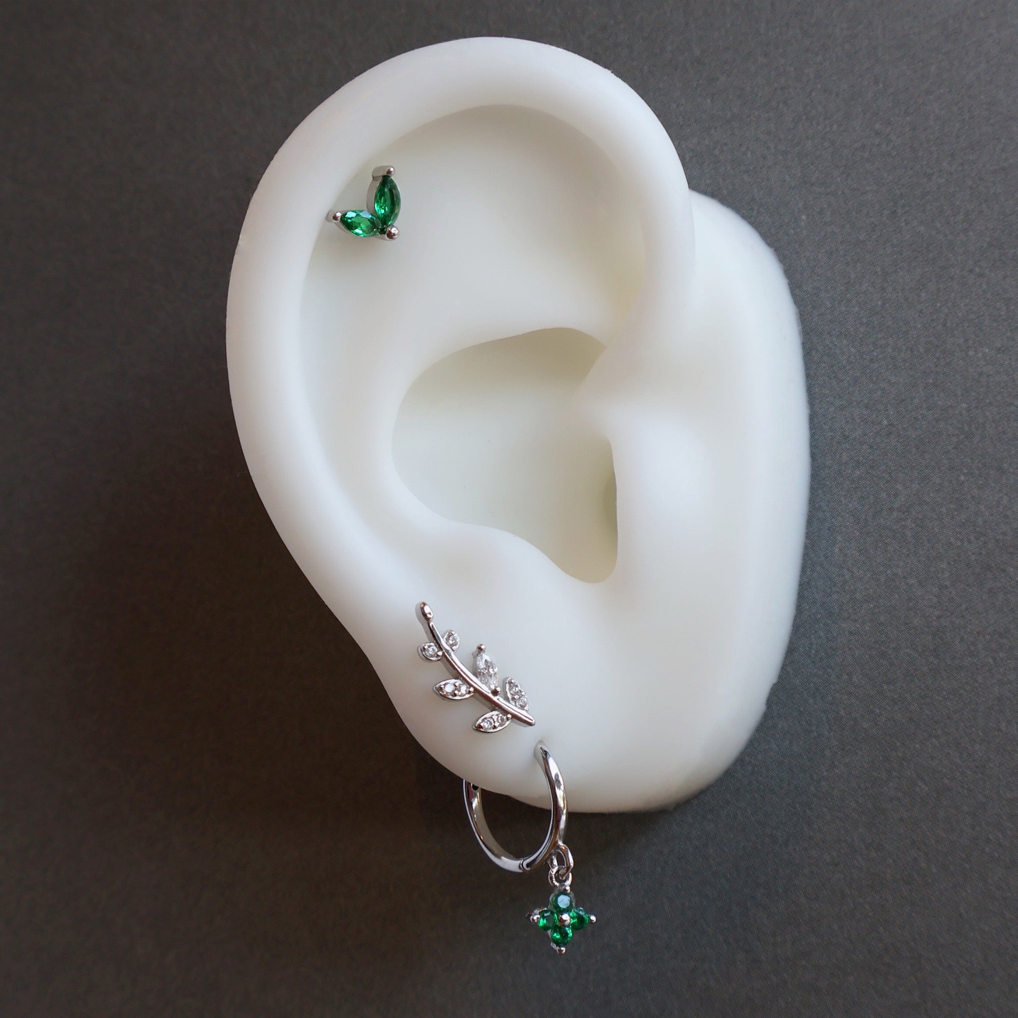 Close-up of a JULE ear piercing stack on a silicone model, showcasing a green clover dangle hoop on the lobe, a silver leaf climber, and a green leaf helix stud.