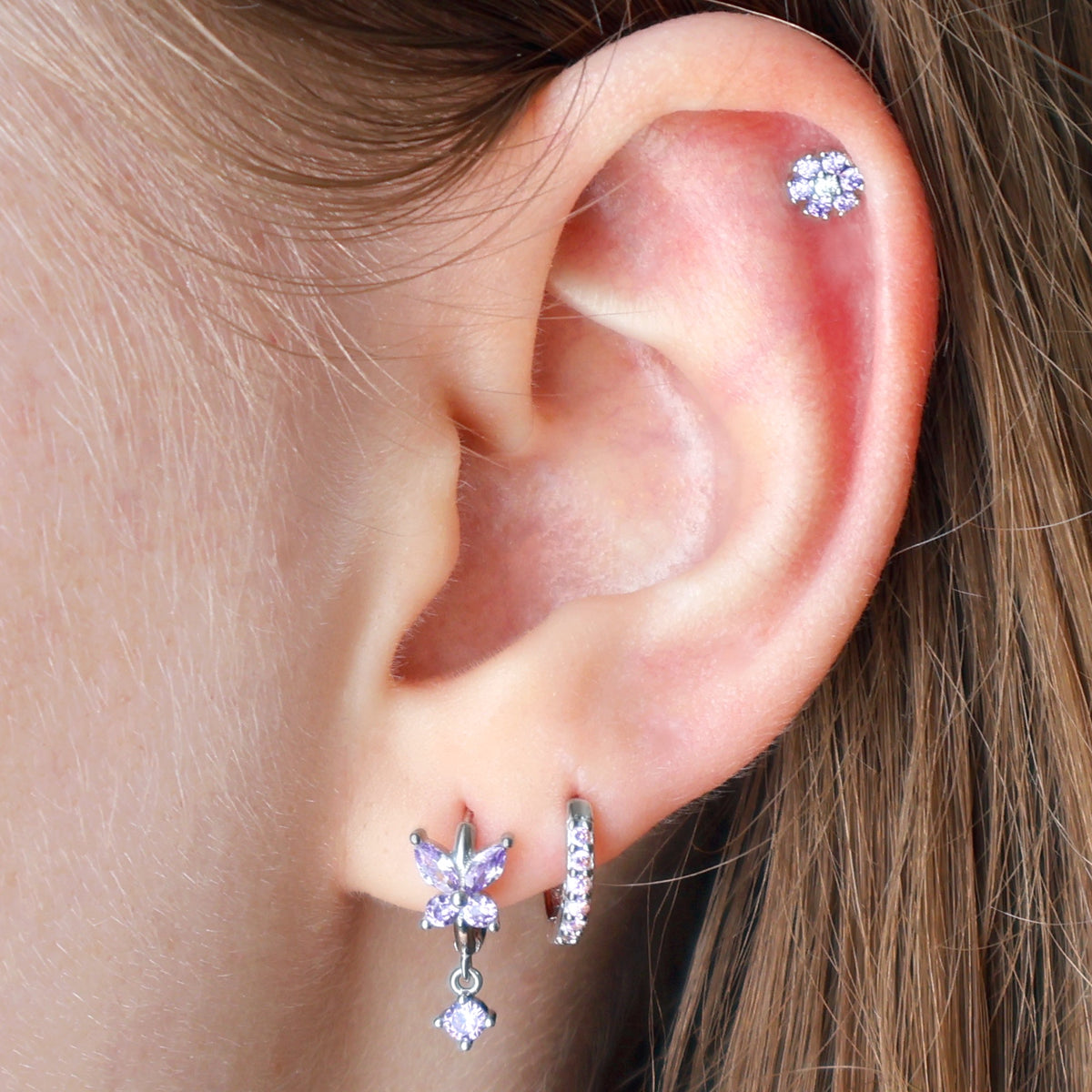 A delicate silver ear piercing stack worn on an ear, featuring a purple crystal butterfly dangle hoop, a matching paved huggie on the lobe, and a sparkling purple flower stud on the helix by JULE.