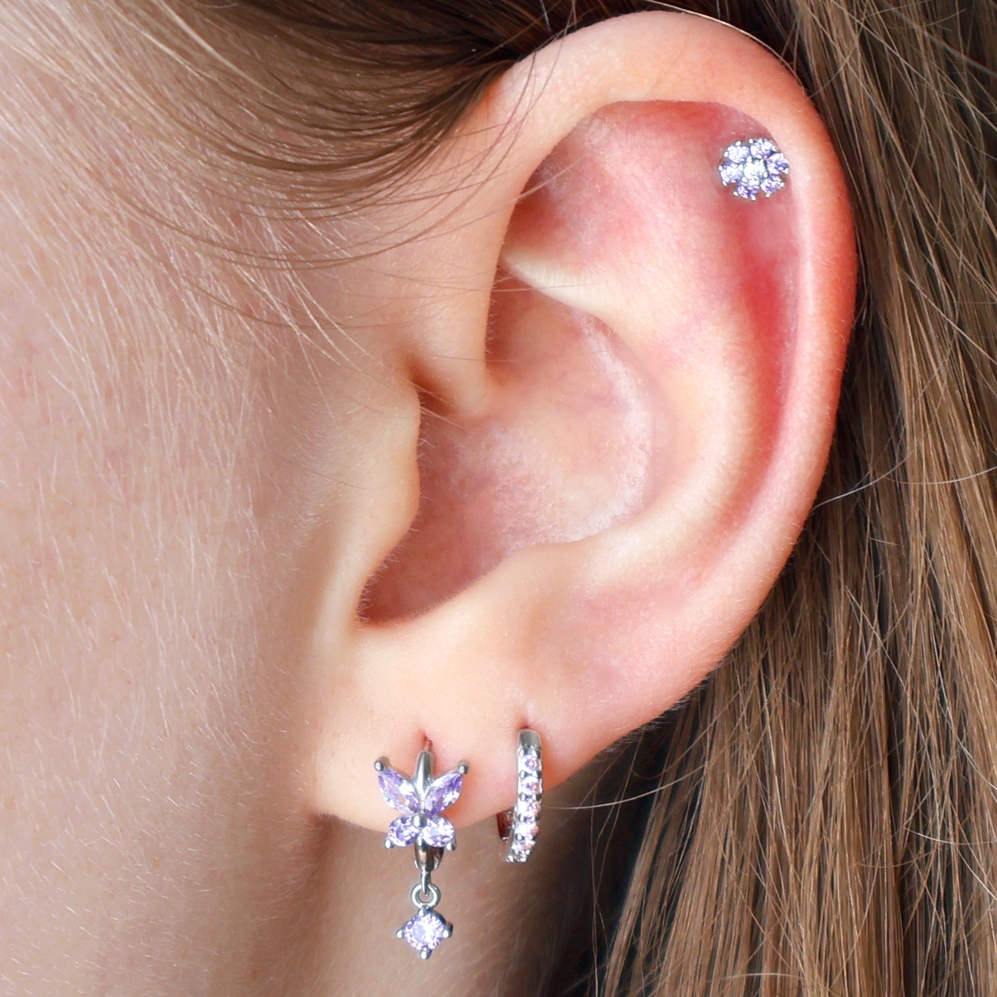 A delicate silver ear piercing stack worn on an ear, featuring a purple crystal butterfly dangle hoop, a matching paved huggie on the lobe, and a sparkling purple flower stud on the helix by JULE.