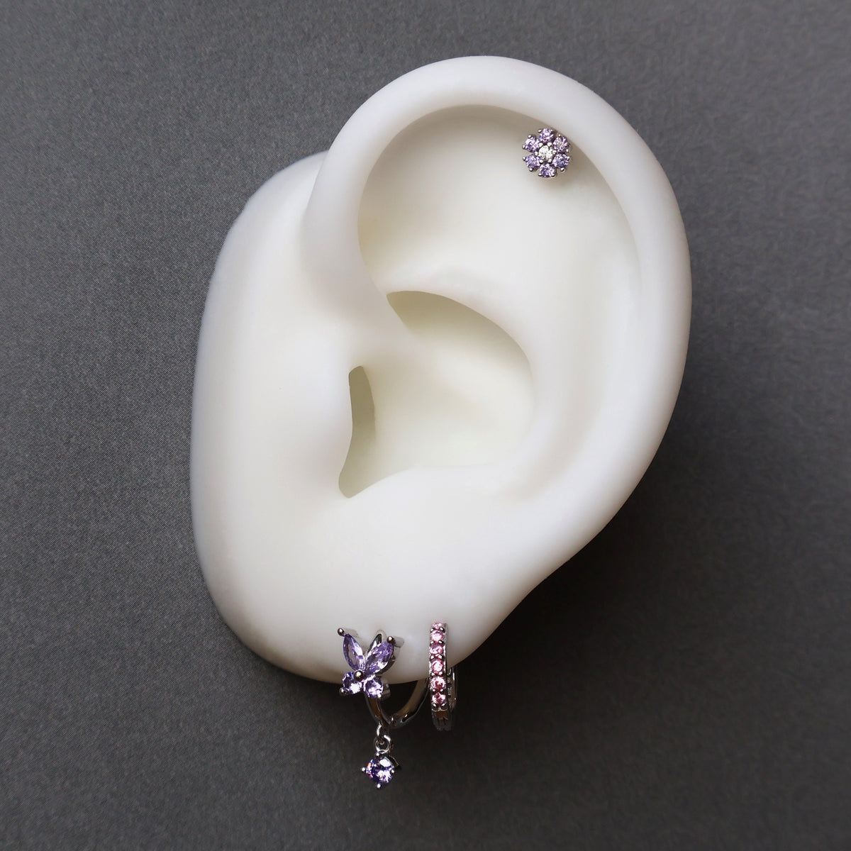 Silver ear piercing stack on a model showcasing a purple crystal butterfly dangle hoop, a matching paved huggie on the lobe, and a purple floral stud on the helix by JULE.