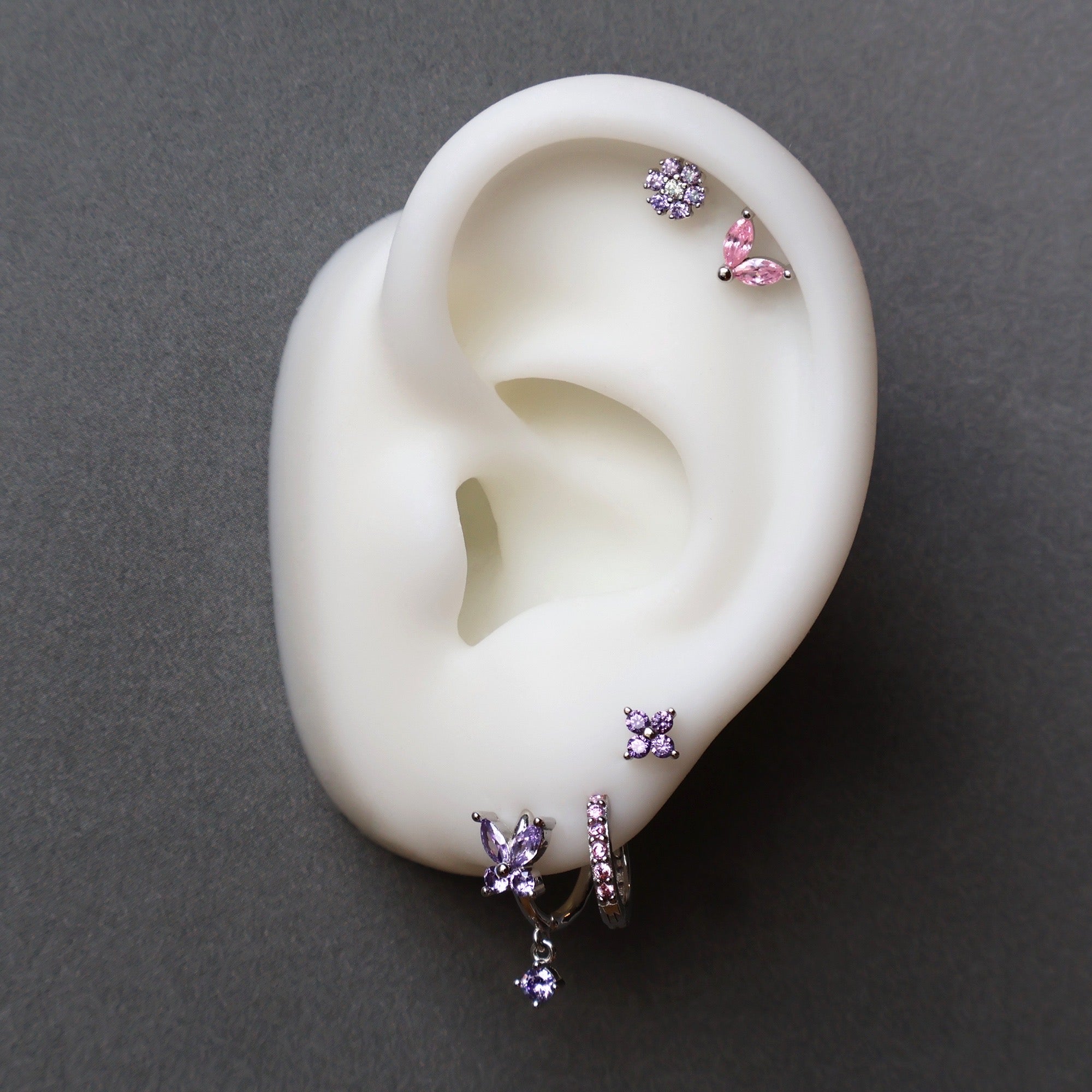 A curated silver ear piercing stack on a white display model by JULE, featuring a purple gemstone butterfly dangle, a paved huggie, a small flower stud on the lobe, and a double helix with pink and purple floral studs.