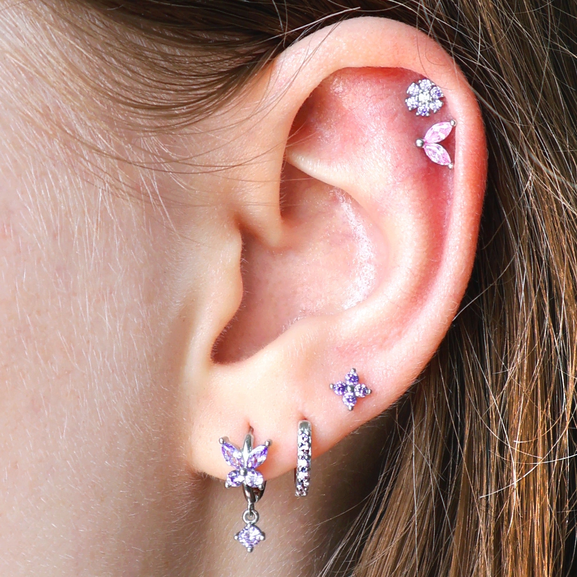 A woman wearing an elaborate JULE silver ear piercing stack, featuring a purple butterfly dangle hoop, a paved huggie, a lobe flower stud, and a beautiful double helix with pink and purple gemstone studs.