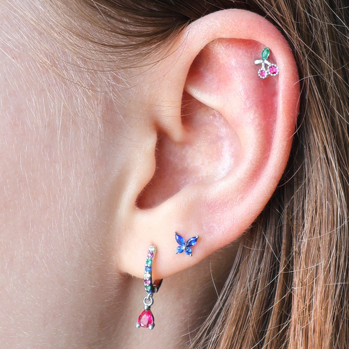 A woman wearing a playful JULE ear piercing stack with a colorful gemstone dangle huggie, a blue butterfly stud on the lobe, and a helix featuring a cherry.