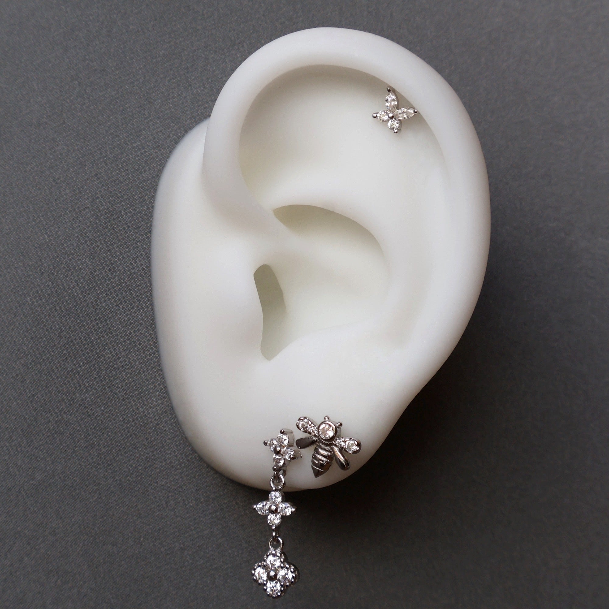 Nature-inspired silver ear piercing stack on a model, showcasing a crystal dangle stud, a sparkling bee piercing, and a delicate floral helix stud.
