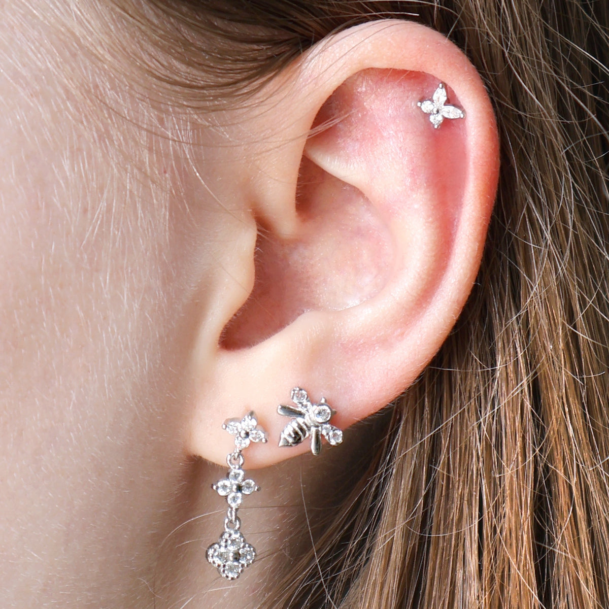 A styled silver ear piercing stack by JULE on a real ear, showing a crystal floral dangle stud, a delicate bee piercing on the lobe, and a small flower stud on the helix.