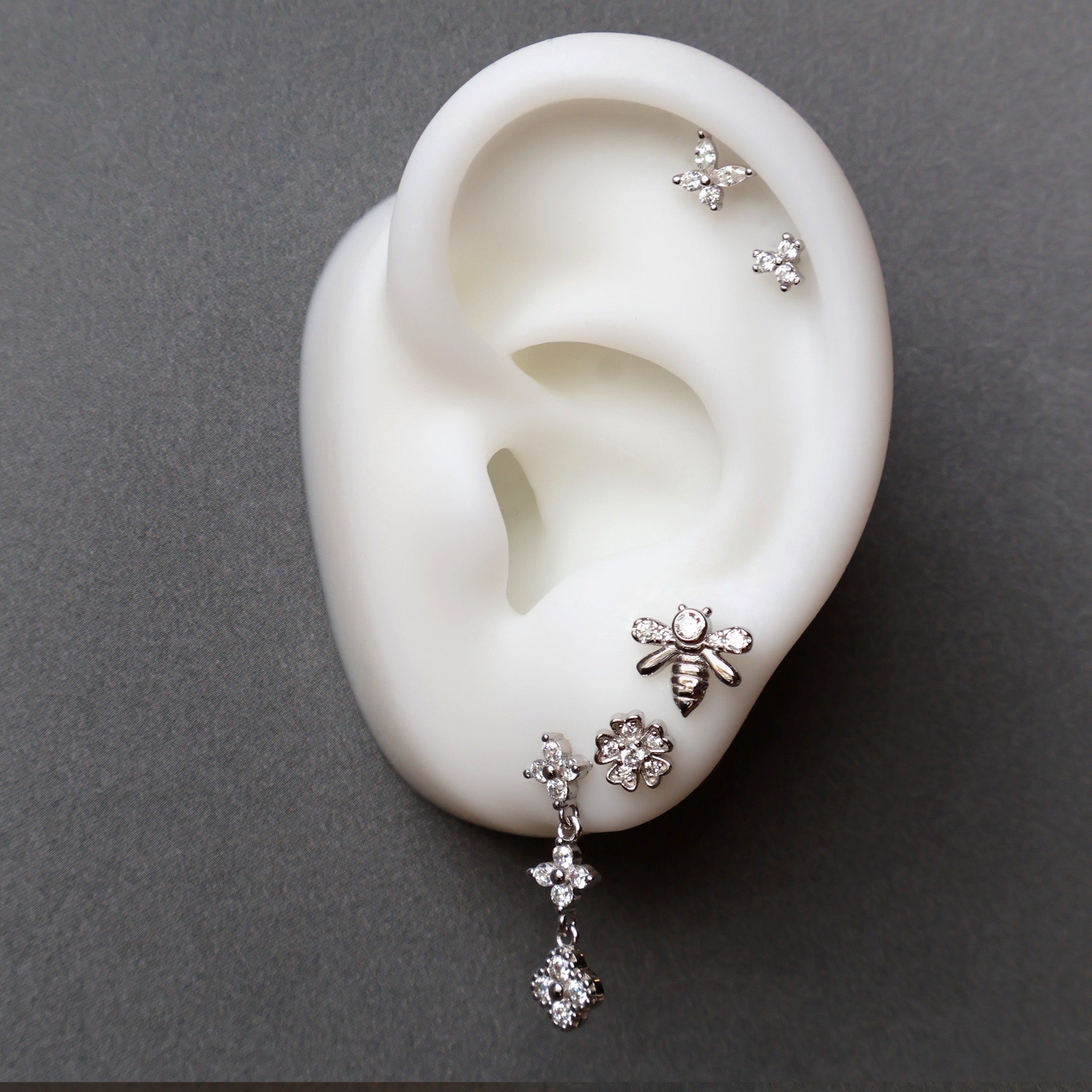 An elaborate silver ear piercing stack by JULE on a display model, featuring a dangle stud, a flower and bee on the lobes, and a matching double floral helix.