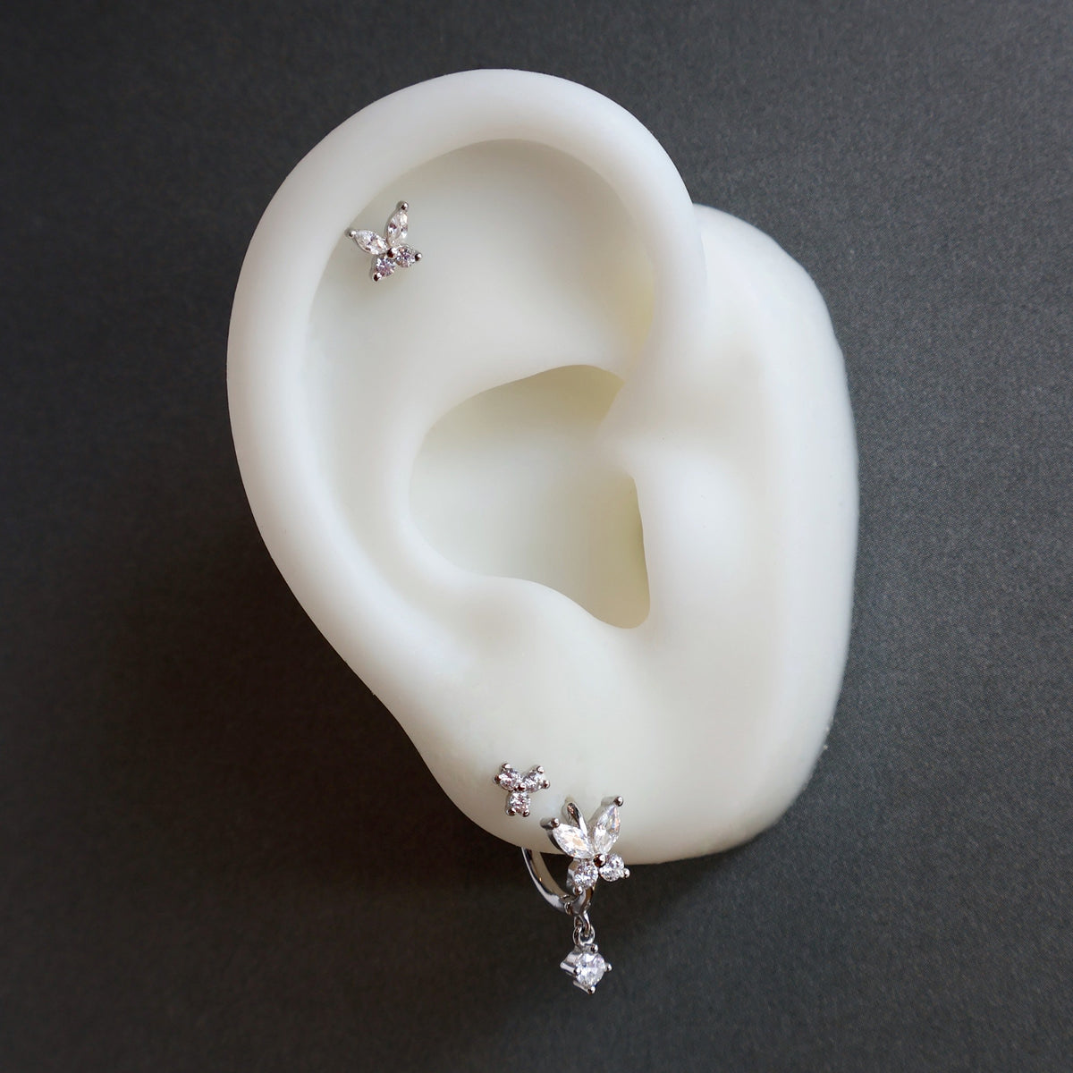 Nature-inspired silver ear piercing stack on a display model by JULE, highlighting a crystal butterfly dangle hoop, a small triple crystal cluster stud on the lobe, and a sparkling butterfly helix piercing.