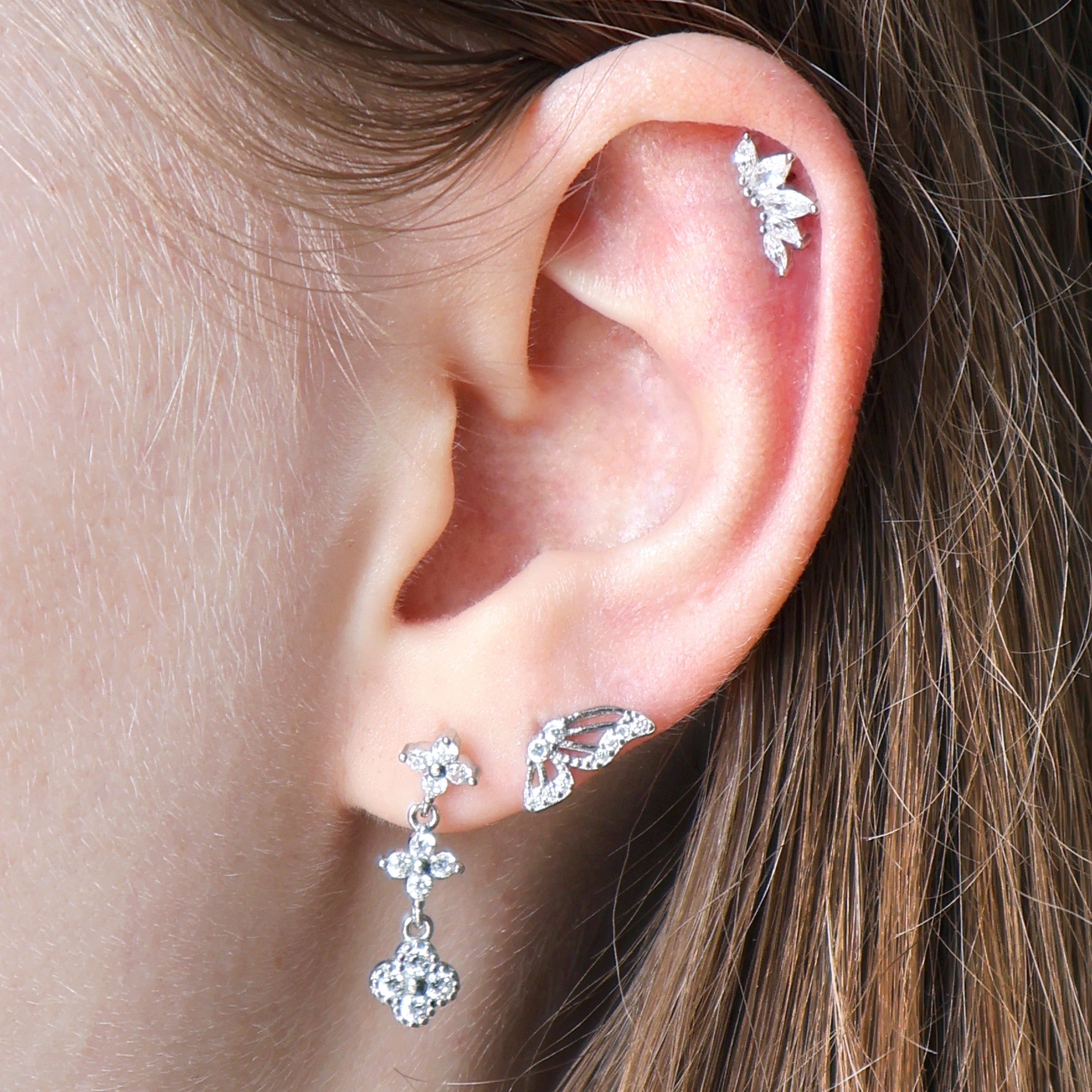 Nature-inspired silver ear piercing stack worn on an ear, featuring a crystal dangle stud, a sparkling butterfly wing on the second lobe, and a leaf crawler on the helix.