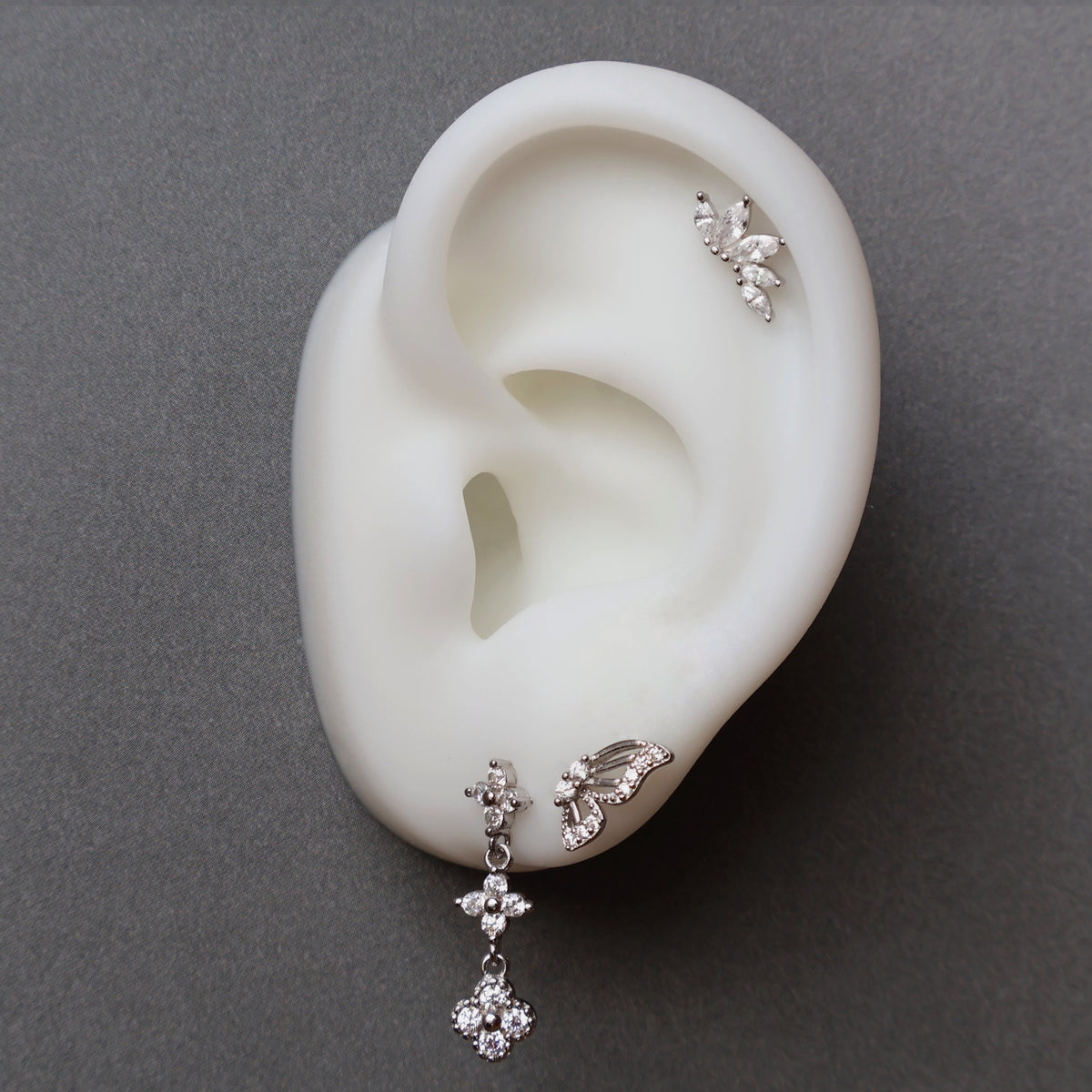 A close-up of a JULE ear piercing stack on a silicone ear, featuring a dangle floral stud, a butterfly wing, and a sparkling marquise leaf crawler on the cartilage.