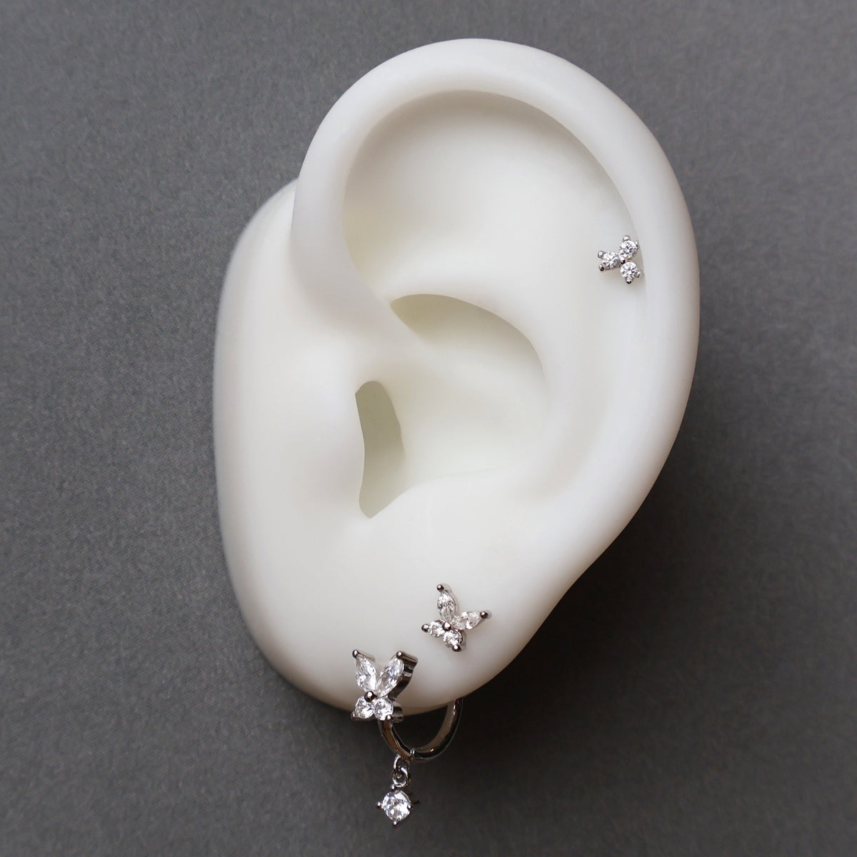 A curated silver ear piercing stack on a white display model by JULE, featuring a clear crystal butterfly dangle hoop, a matching small butterfly stud on the lobe, and a delicate crystal cluster piercing on the helix.