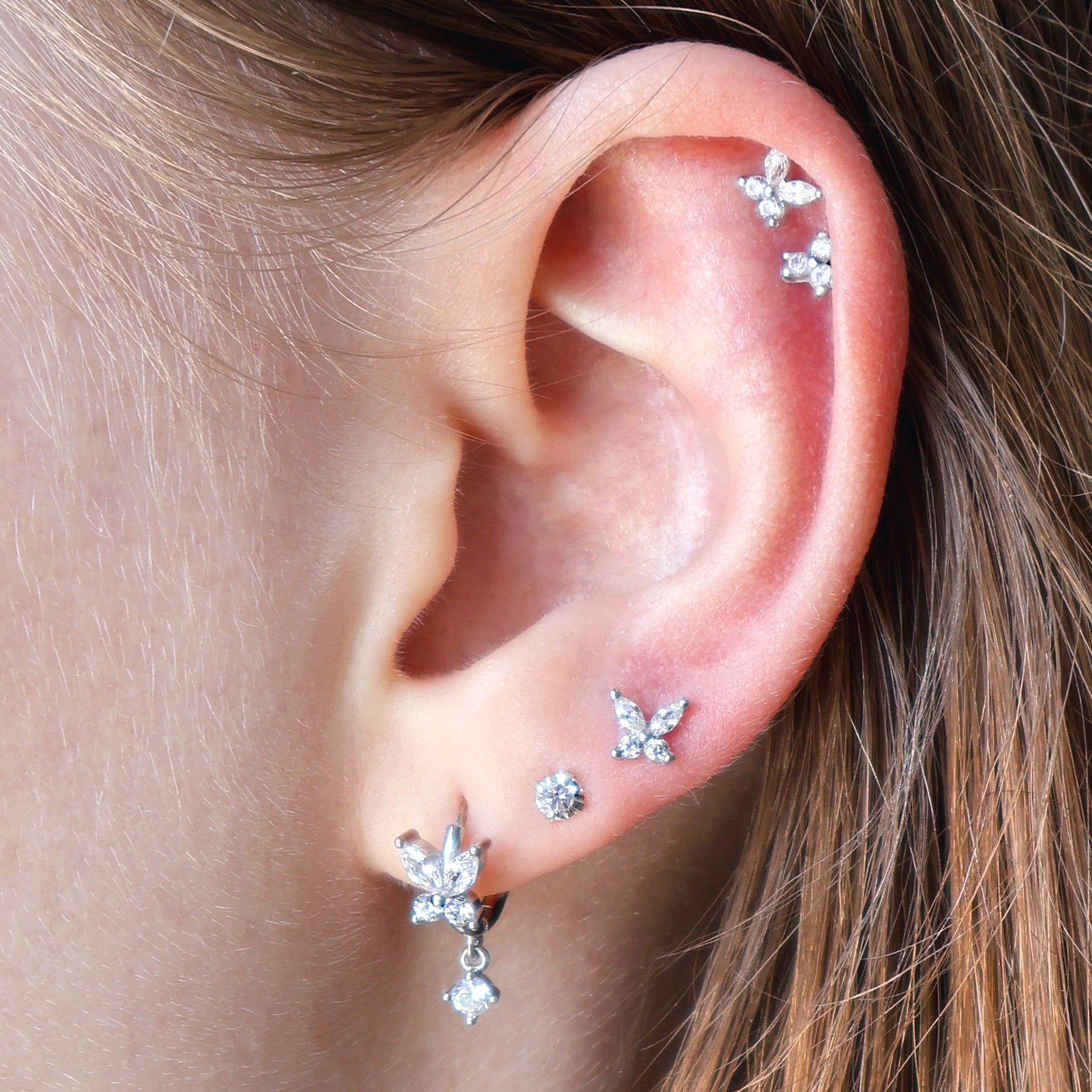 A detailed view of a styled JULE ear piercing stack on a real ear, featuring a crystal butterfly dangle hoop, multiple delicate lobe studs, and a sparkling double butterfly helix.