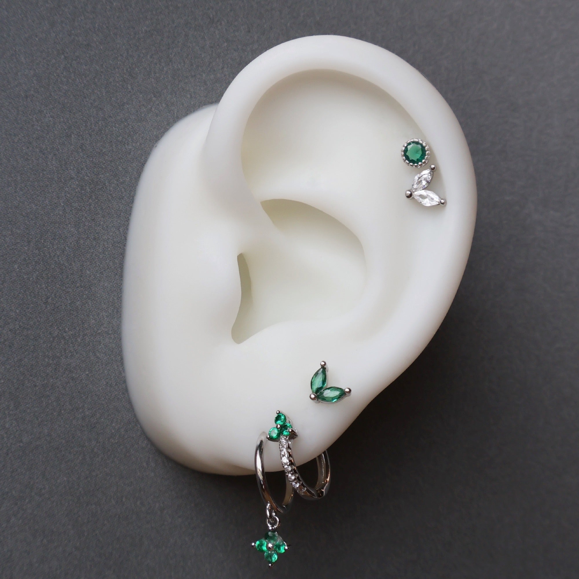 A curated silver ear piercing stack on a white display model by JULE, featuring a green clover dangle hoop, a paved huggie, a green leaf stud on the upper lobe, and a double helix piercing with round and leaf-shaped studs.