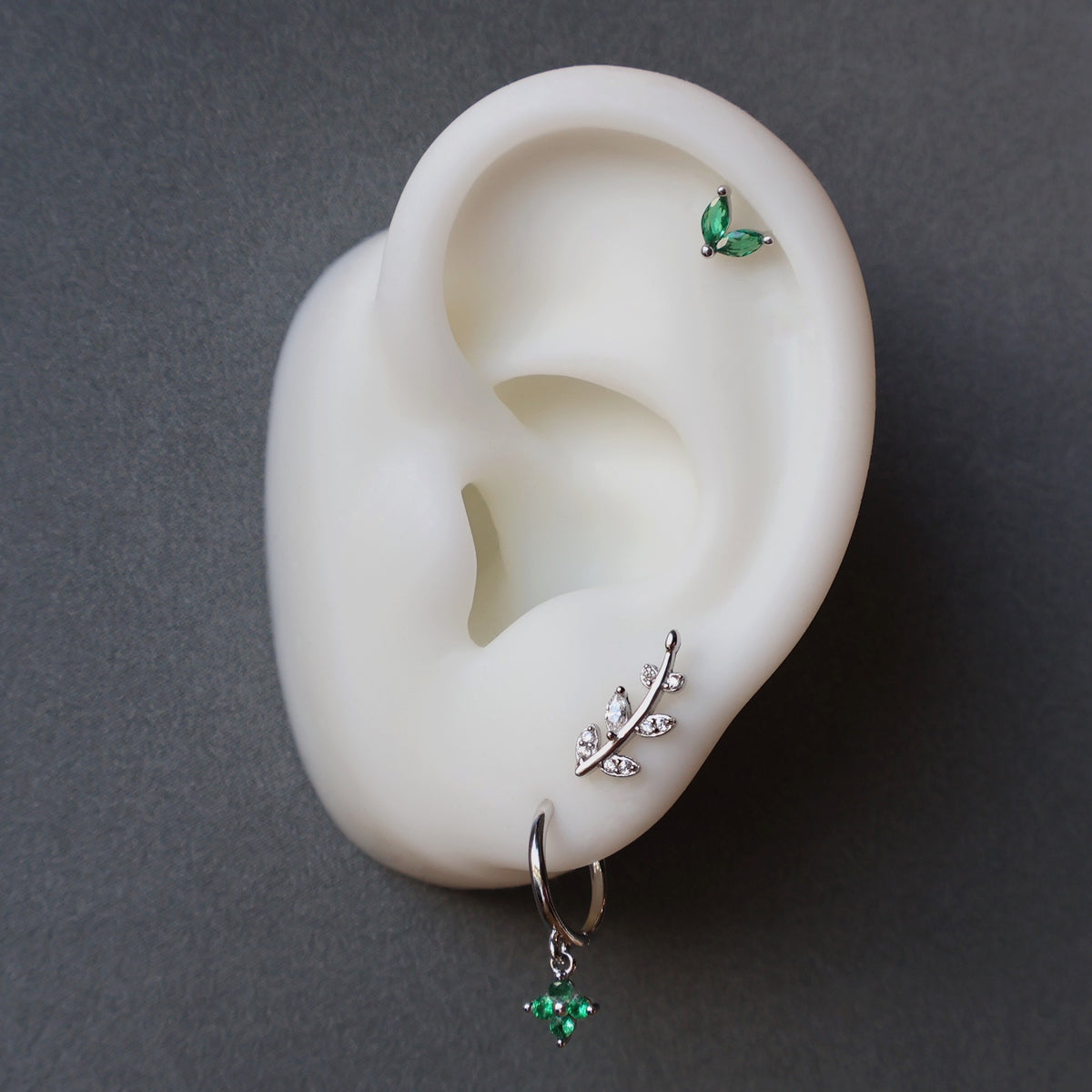 Nature-inspired silver ear piercing stack on a model, featuring a green dangle hoop, a silver leaf crawler, and a two-leaf green gemstone helix stud.
