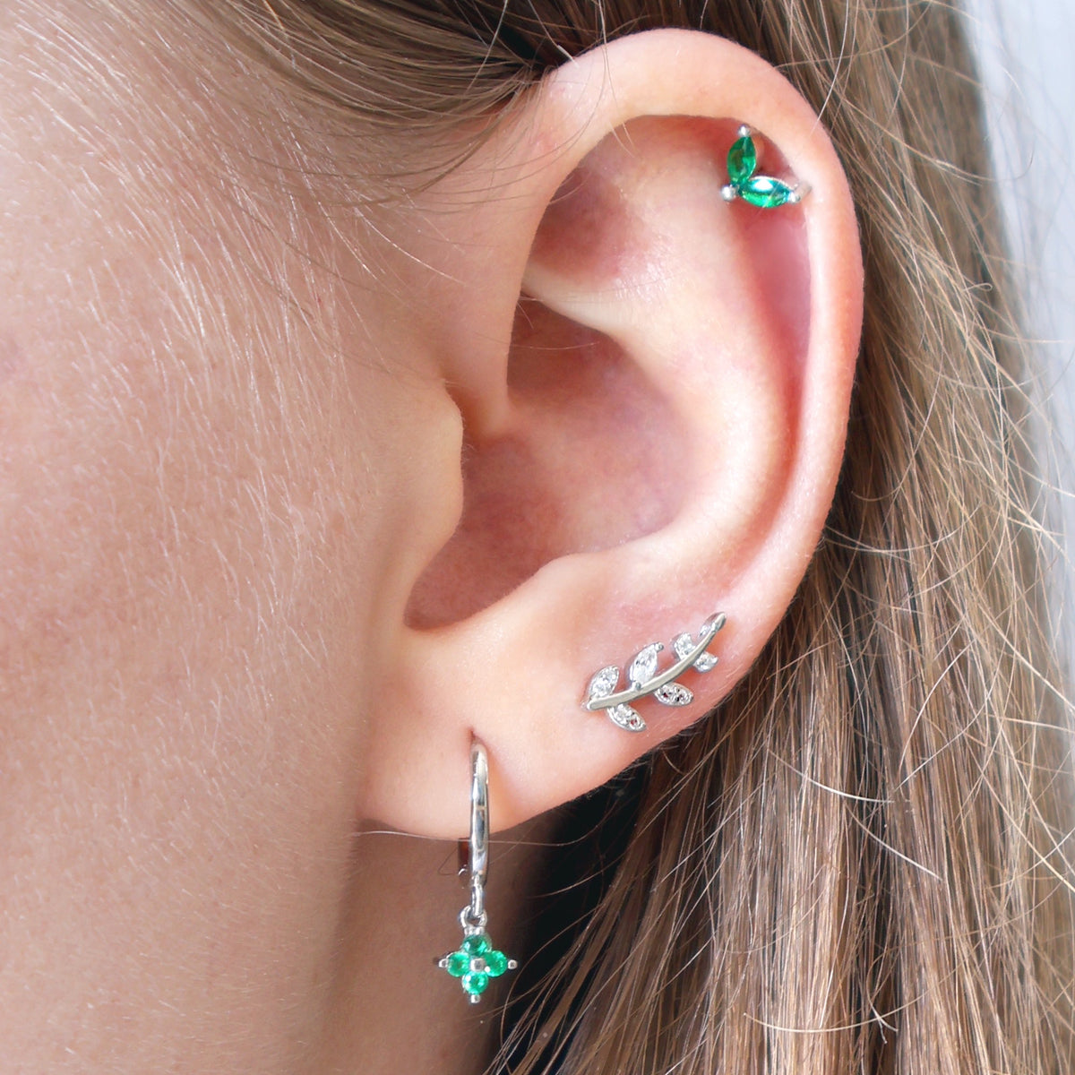 Curated silver ear piercing stack by JULE worn on an ear, featuring a delicate green dangle hoop, a silver leaf stud, and a matching green gemstone helix piercing.
