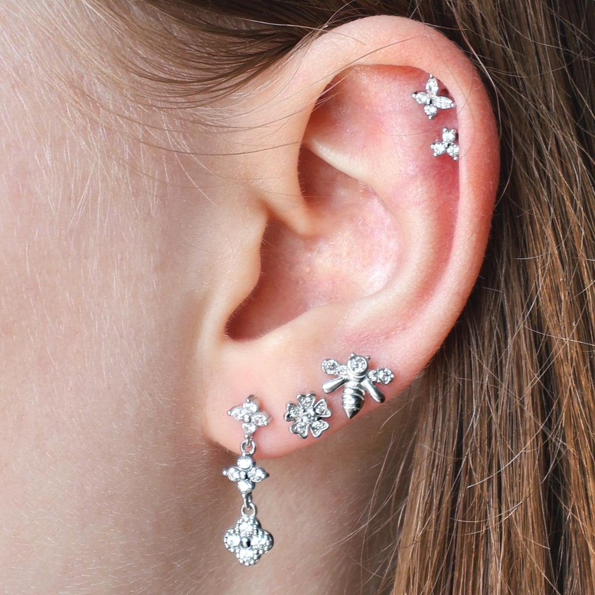 A woman wearing a beautiful silver ear piercing stack by JULE, featuring a crystal dangle stud, floral and bee lobe piercings, and a delicate double floral helix.