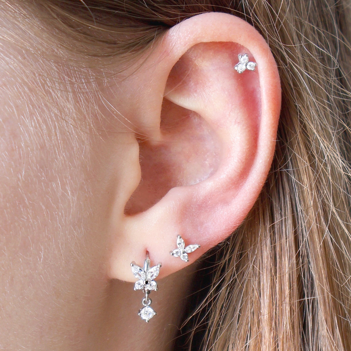 Close-up of a silver ear piercing stack worn on an ear, showcasing a JULE crystal butterfly dangle hoop, a small matching butterfly stud on the lobe, and a tiny crystal cluster piercing on the cartilage.
