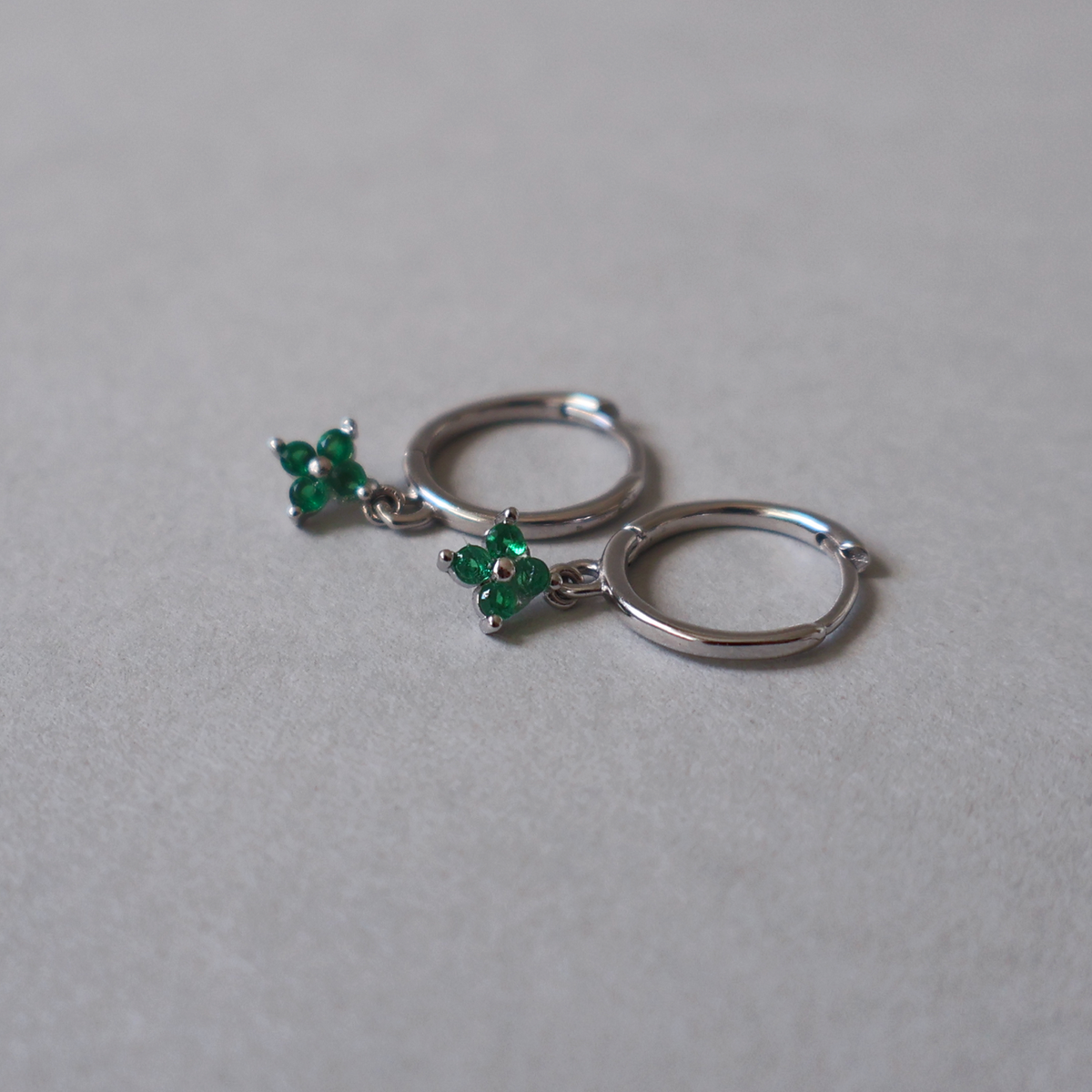 925 sterling silver hoop earrings featuring a dangling green cubic zirconia four-leaf clover design, resting on a light neutral background.
