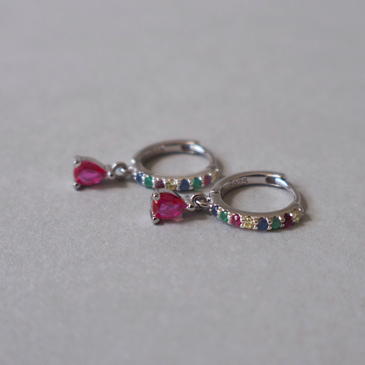 925 sterling silver hoop earrings set with small multicolored cubic zirconia and a bright pink dangling teardrop stone, resting on a light neutral background