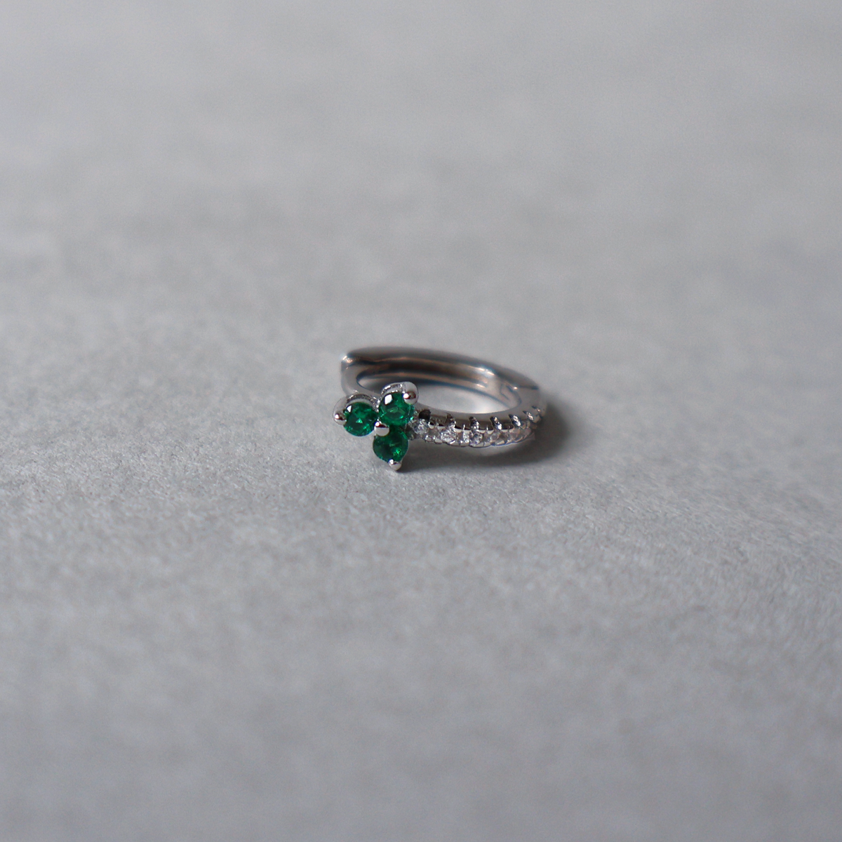 Silver huggie hoop earring set with small clear cubic zirconia along the band and a prominent three-stone shamrock or clover motif made of deep green gemstones. Detailed photo of the jewelry resting on a light neutral background.