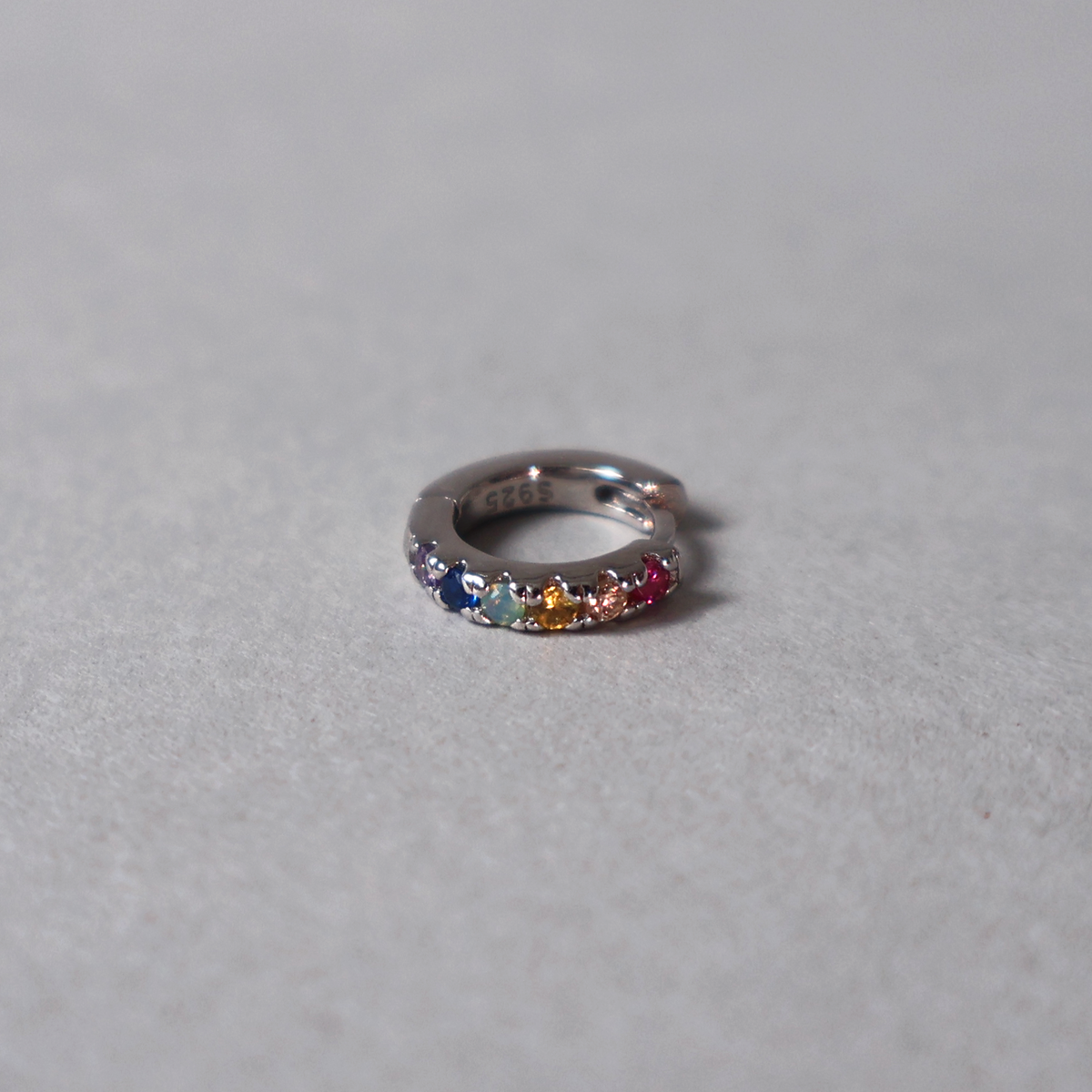 925 sterling silver huggie hoop earring set with a row of multicolored cubic zirconia in a rainbow pattern. Detailed photo of the jewelry resting on a light neutral background.