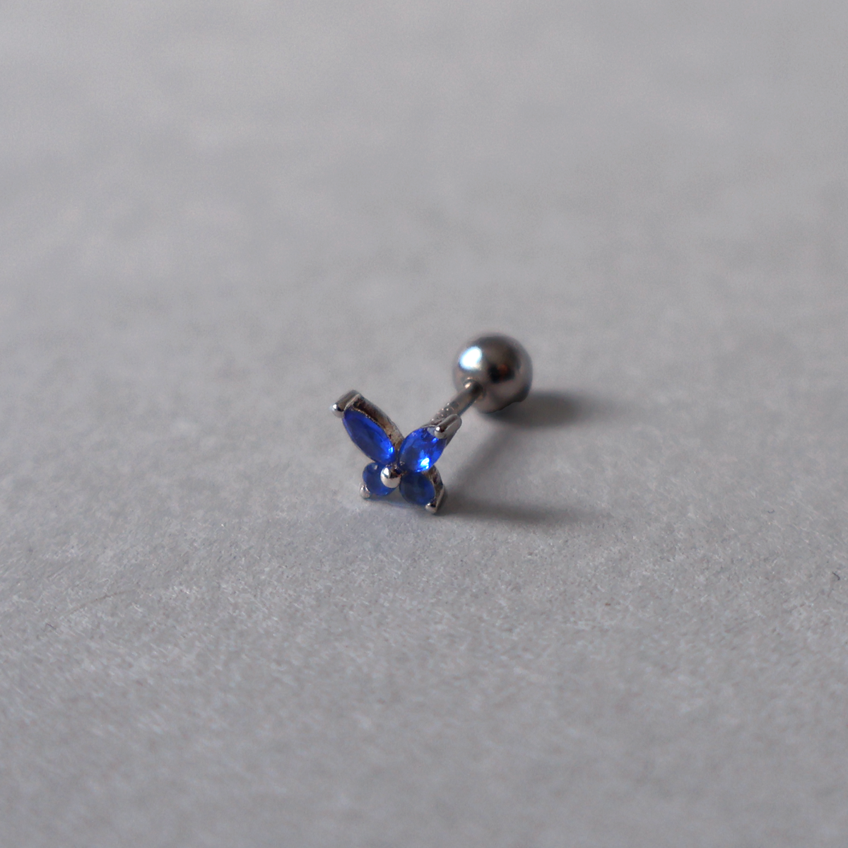Silver piercing stud featuring a delicate butterfly design set with vibrant blue cubic zirconia. Detailed photo of the jewelry resting on a light neutral background.