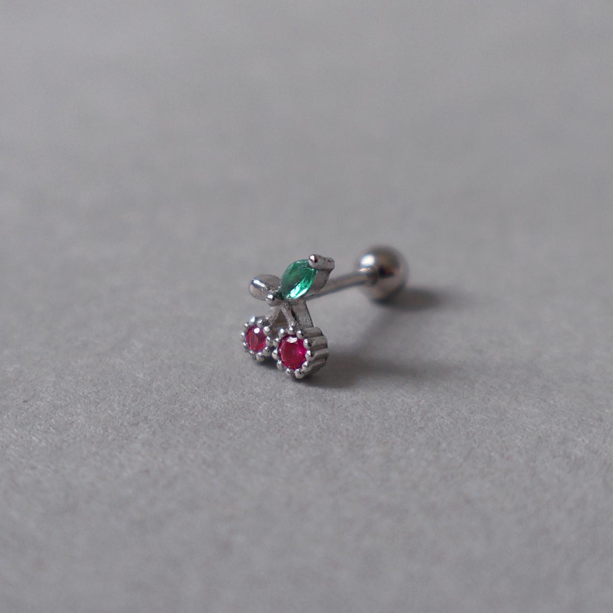 Silver piercing stud featuring a cherry design set with pink and green cubic zirconia. Detailed photo of the jewelry resting on a light neutral background.