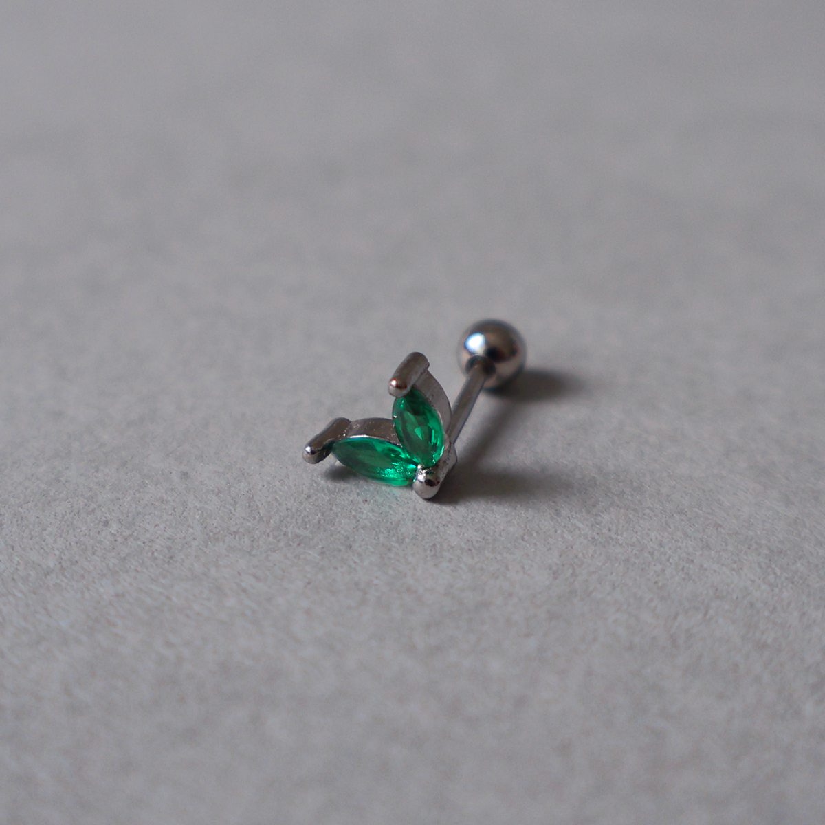 Silver piercing stud featuring a delicate two-leaf design set with emerald green cubic zirconia. Detailed photo of the jewelry resting on a light neutral background.