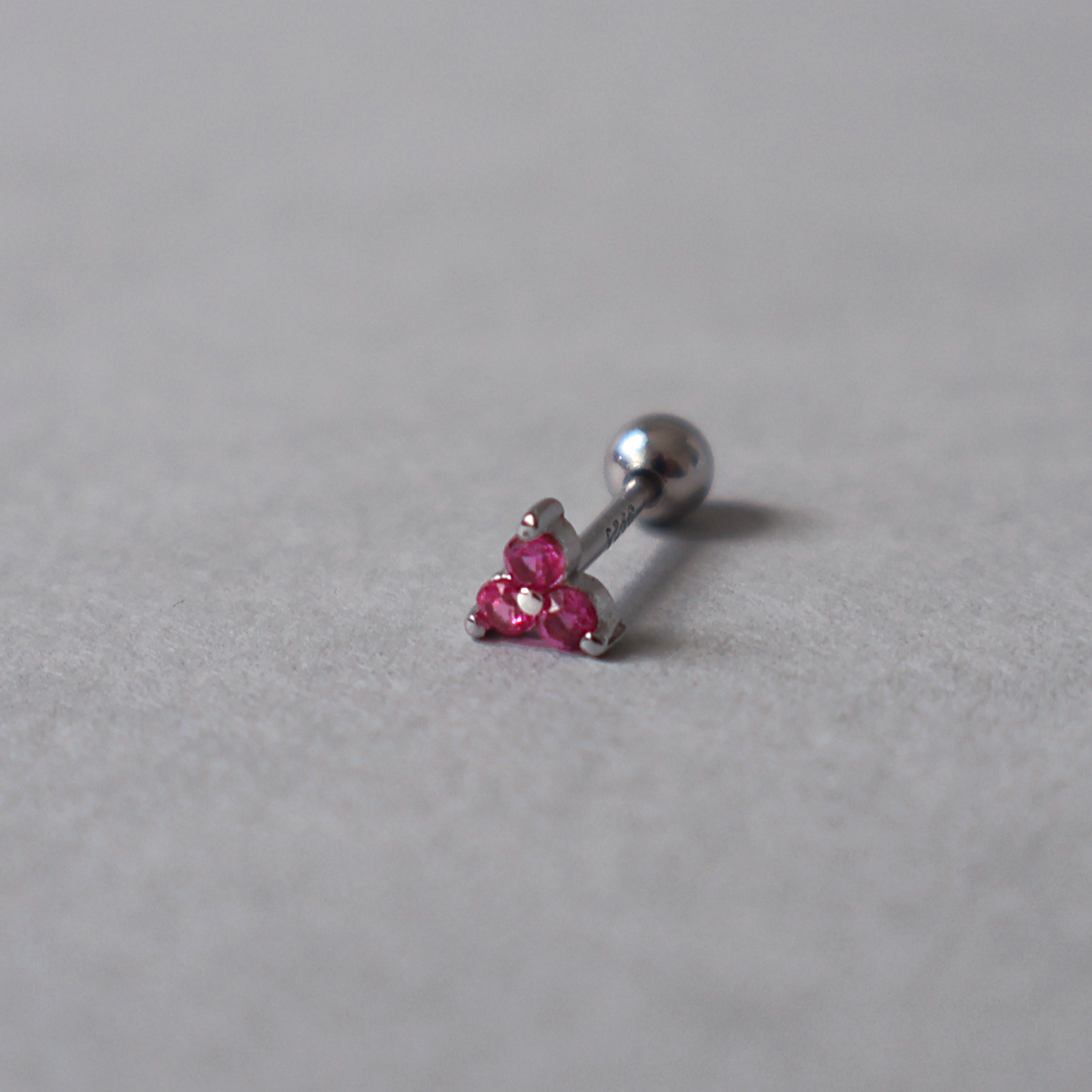 Silver piercing stud featuring a small cluster of three bright pink cubic zirconia stones. Detailed photo of the jewelry resting on a light neutral background.