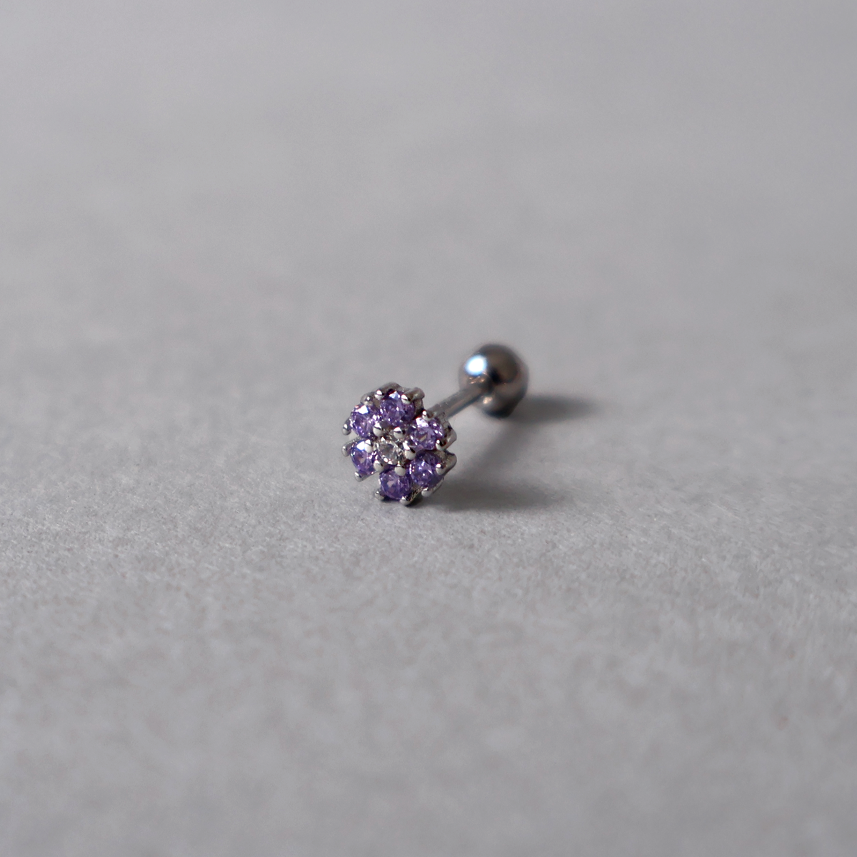 Silver piercing stud featuring a flower design with a clear center cubic zirconia surrounded by purple stones. Detailed photo of the jewelry resting on a light neutral background.