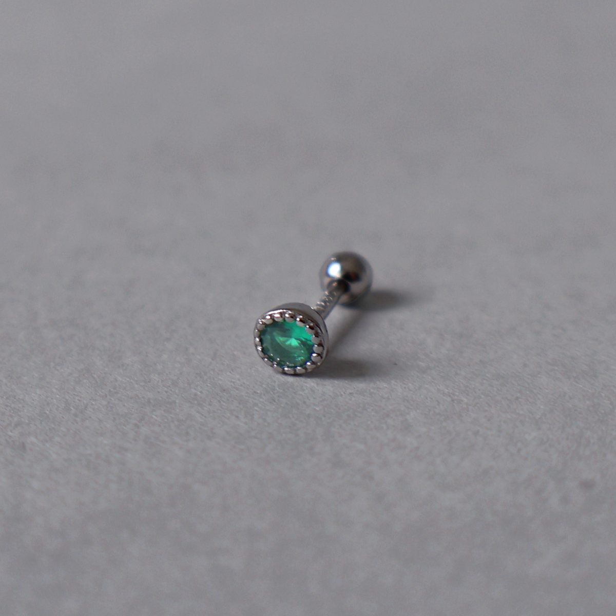 Silver piercing stud featuring a single round emerald green cubic zirconia in a beaded bezel setting. Detailed photo of the jewelry resting on a light neutral background.